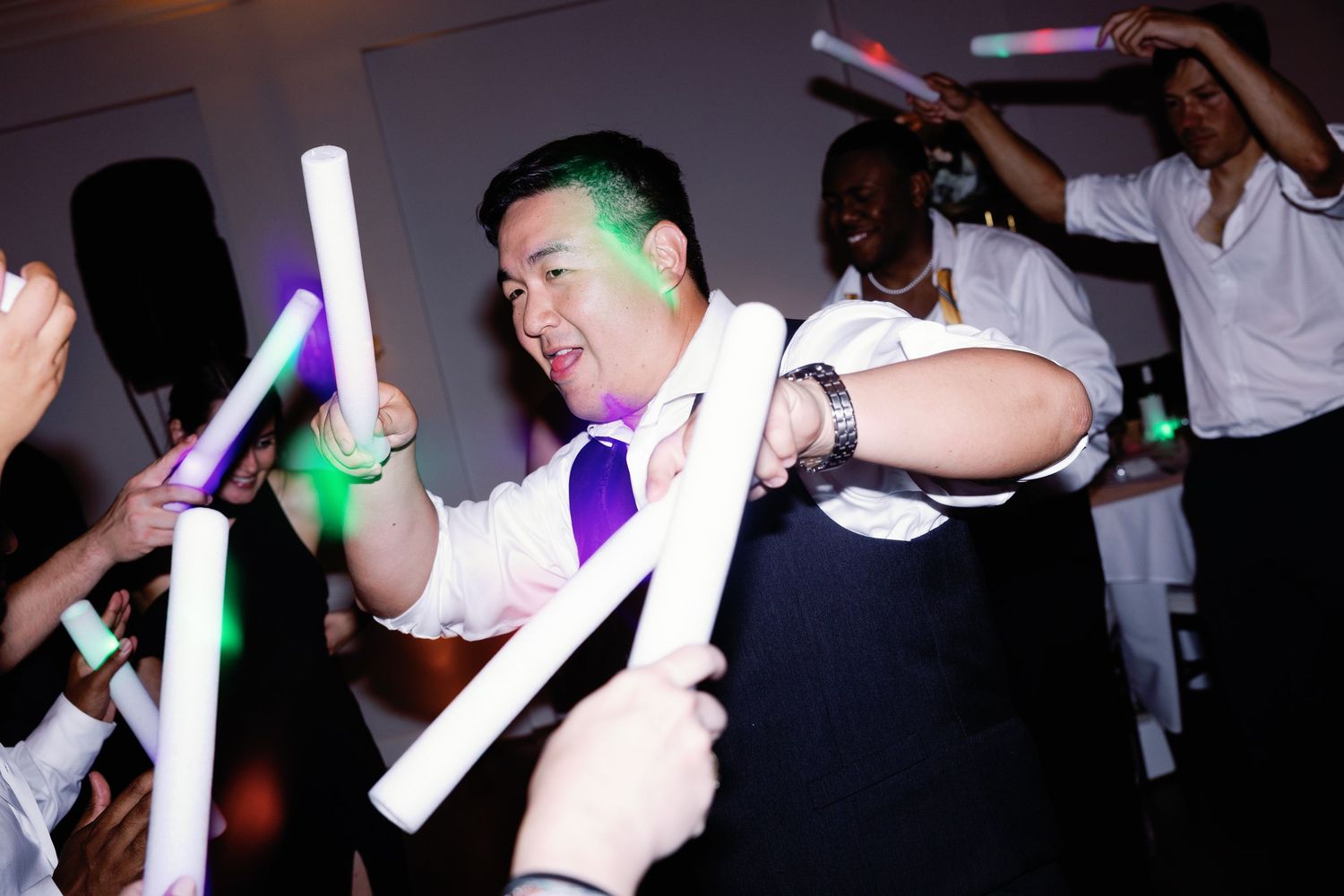 Glowing foam sticks create light trails as wedding guests dance in a darkened reception space.