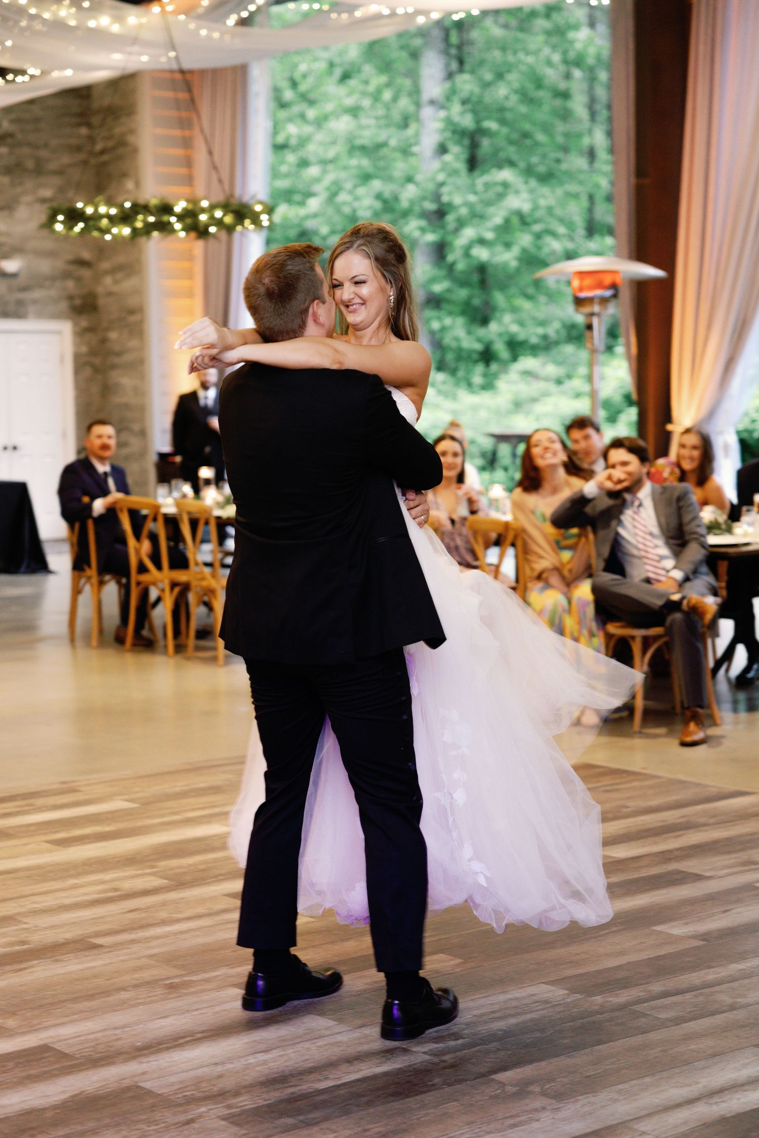 Wedding couple dances on rustic wooden floor with reception venue in background.