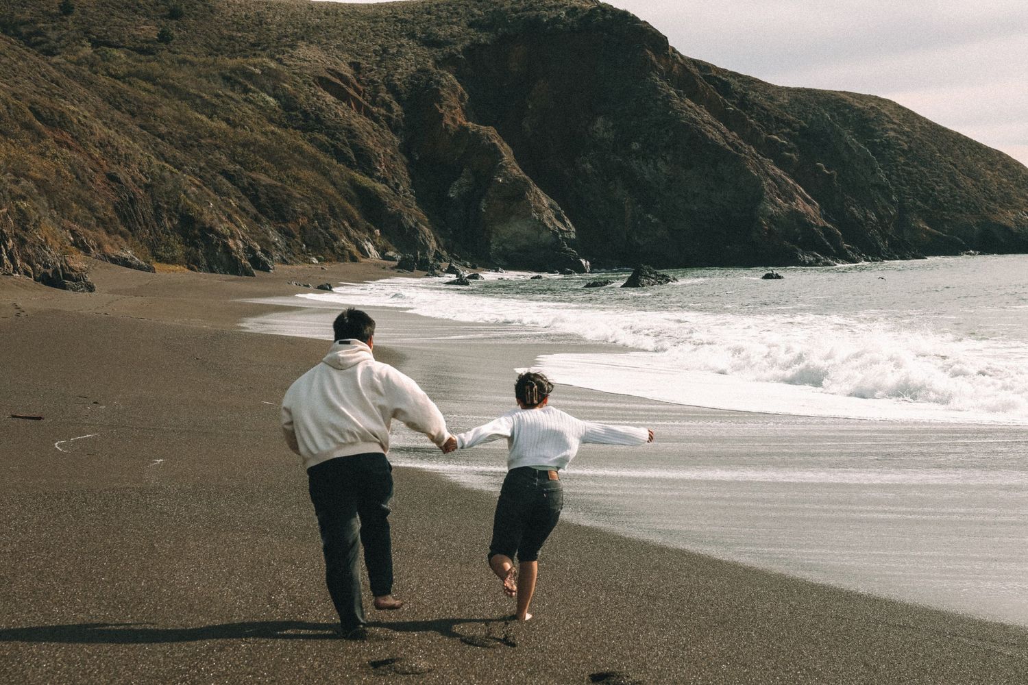 Two people walking along a dramatic black sand beach with cliffs in the distance.