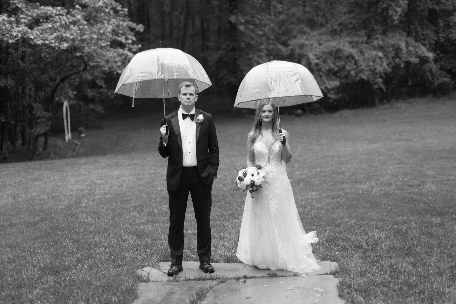 Black and white photo of newlyweds holding clear umbrellas on a rainy wedding day.