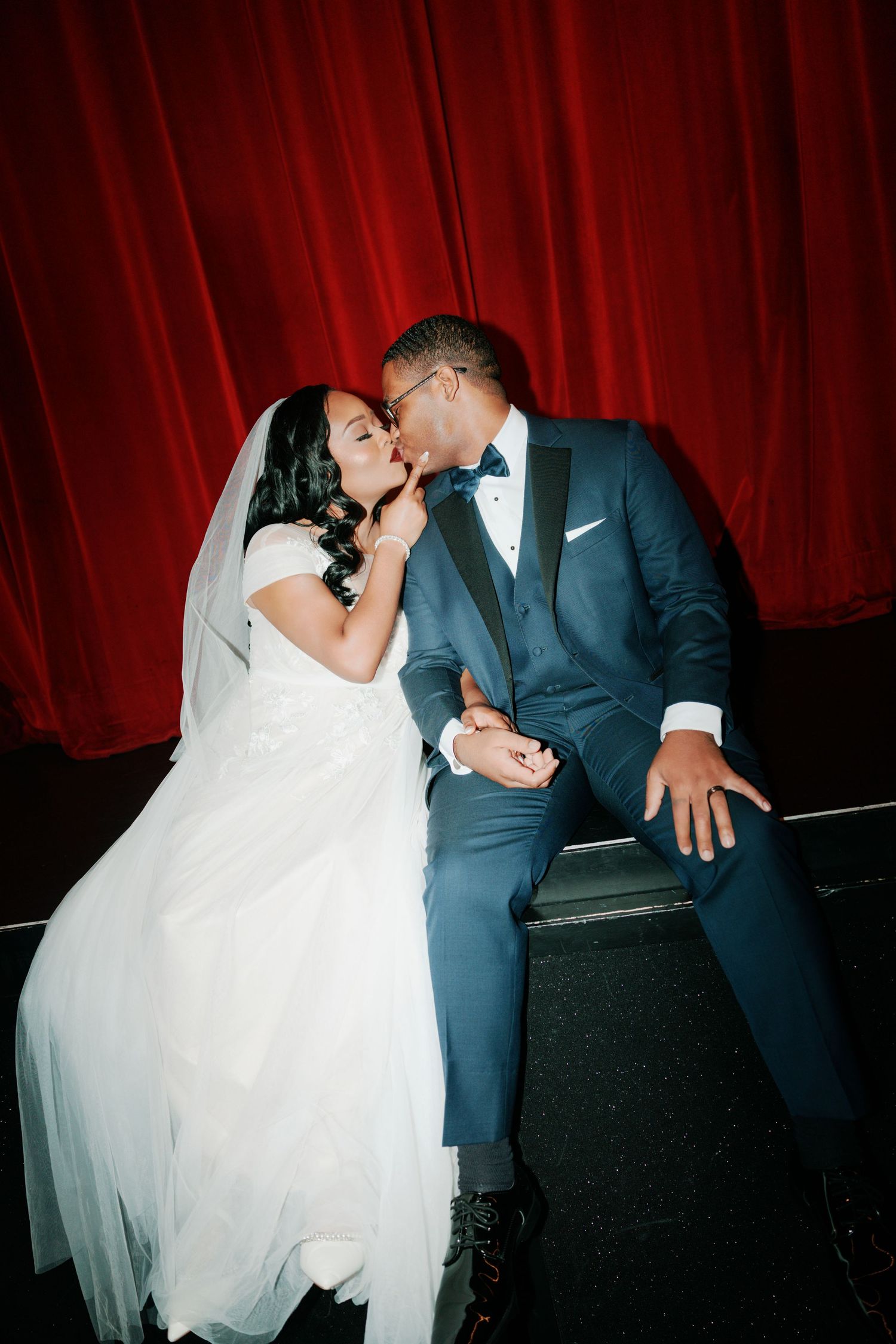 Intimate moment between newlyweds against red theater curtain.