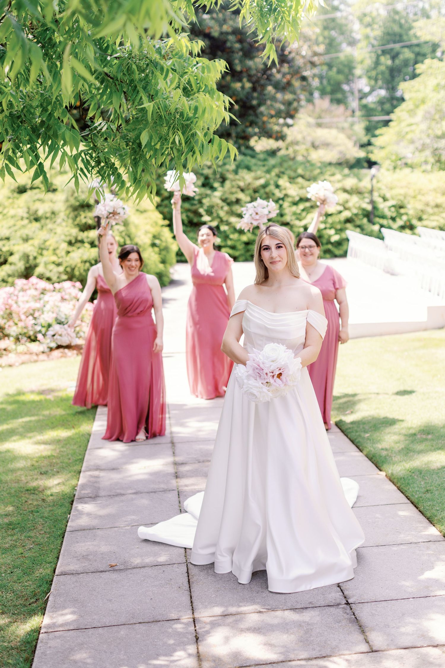 Bridal party in rose-colored dresses walking along a stone pathway under green foliage.