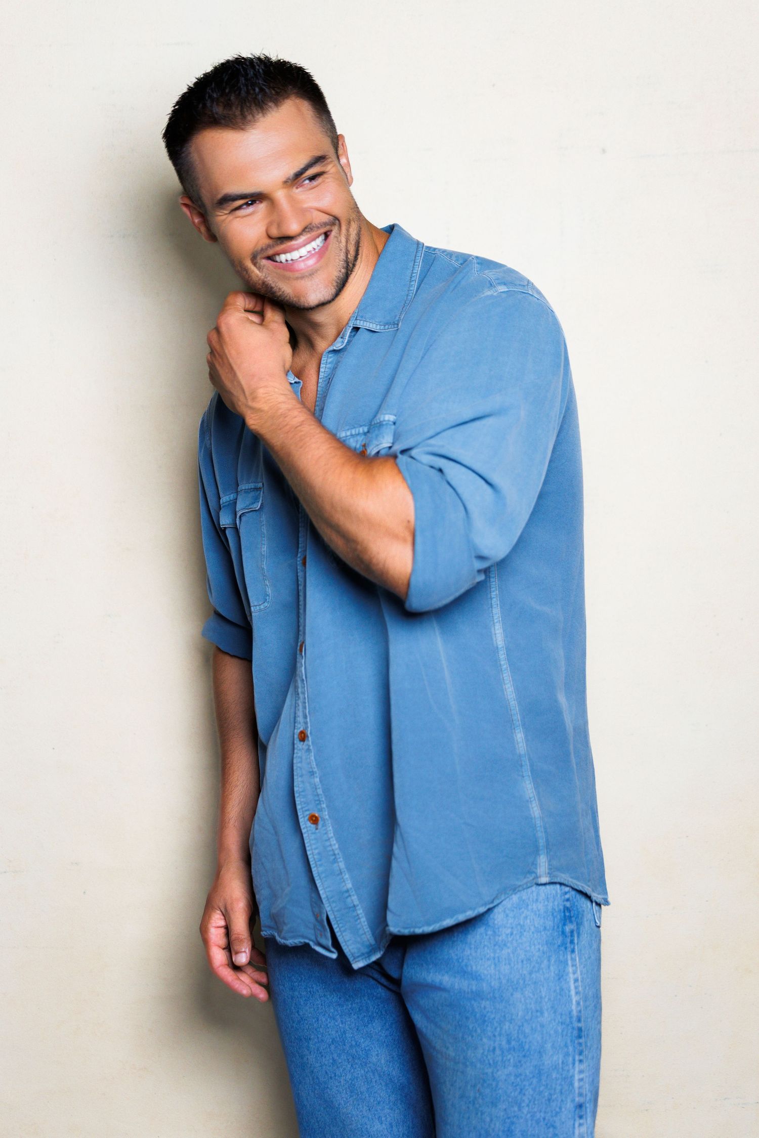 Person in casual blue button-down shirt and jeans poses against light background with warm smile.