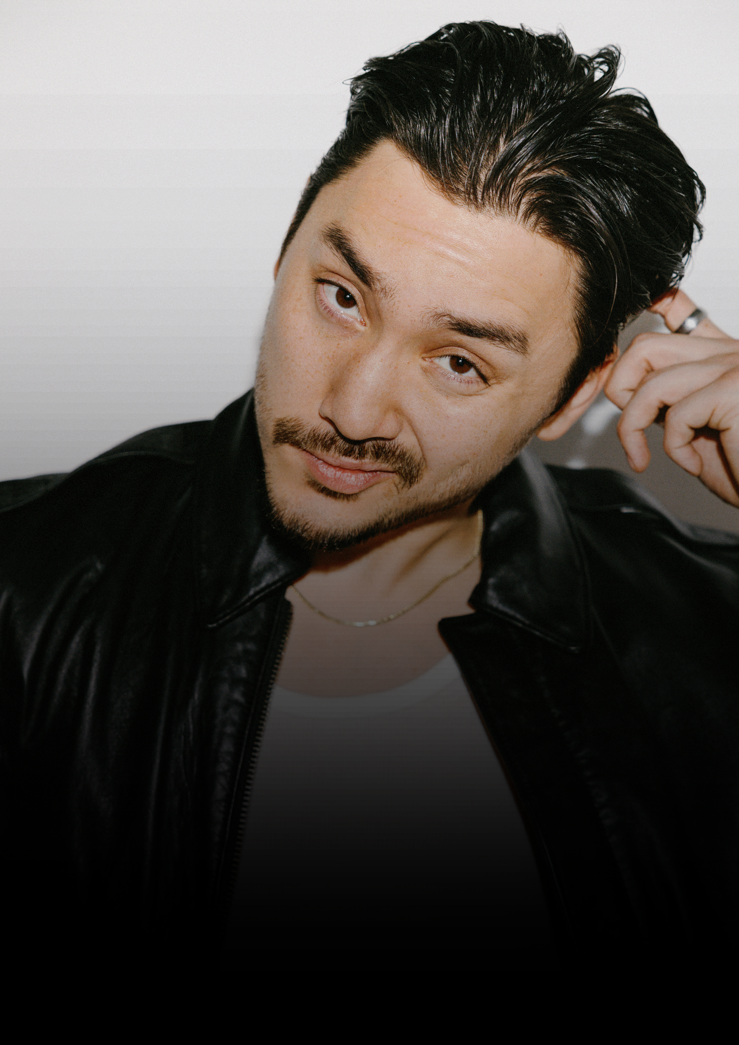 A young man with dark hair in a black leather jacket poses confidently against a light background.