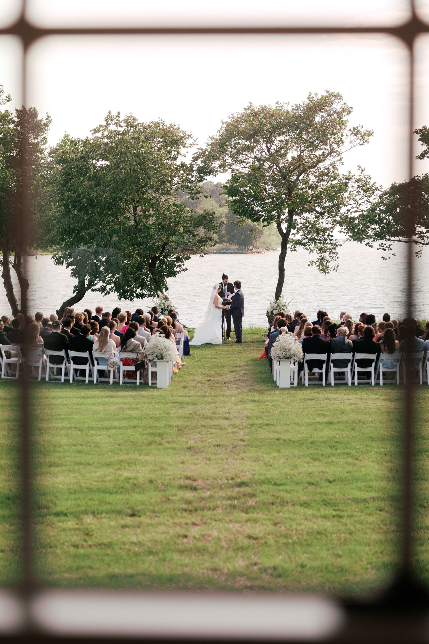 Outdoor wedding ceremony setup by a lake with guests seated in white chairs.