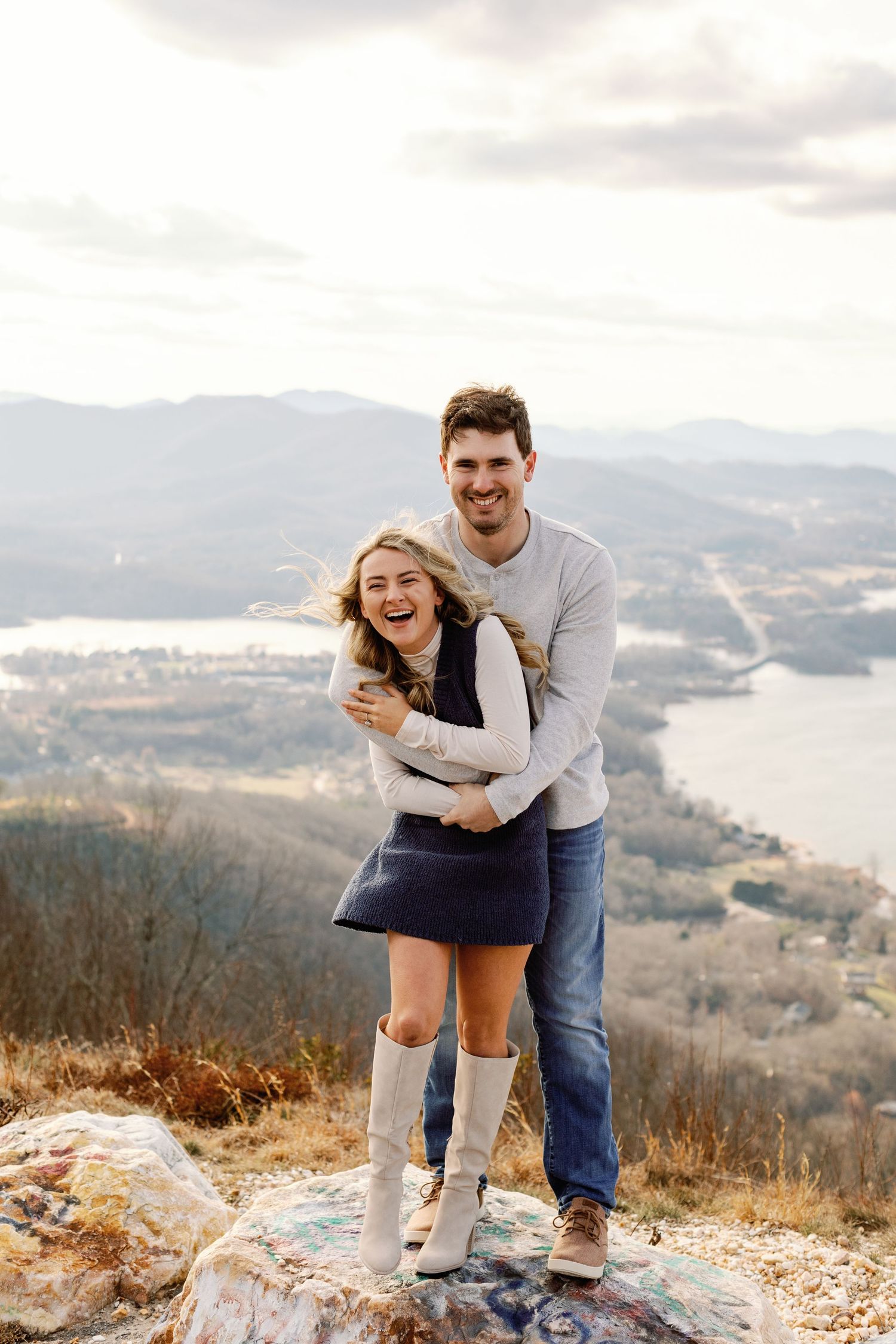 Playful couple embracing on a mountain overlook with a scenic lake view in the background.