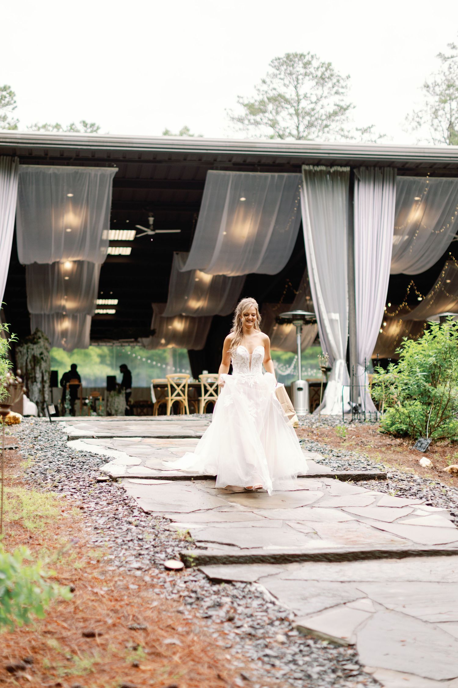 A bride in a flowing white dress stands in front of an outdoor venue with draped white curtains and elegant lighting.