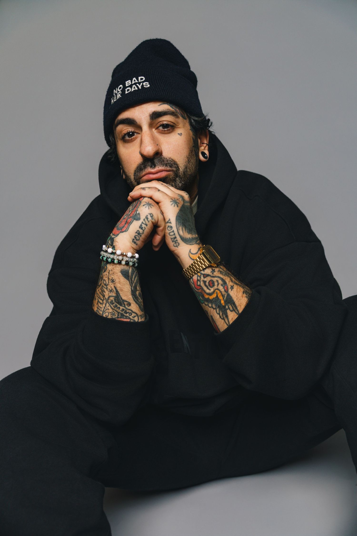 A person wearing a black hoodie and beanie poses against a gray backdrop, showing heavily tattooed hands and arms.