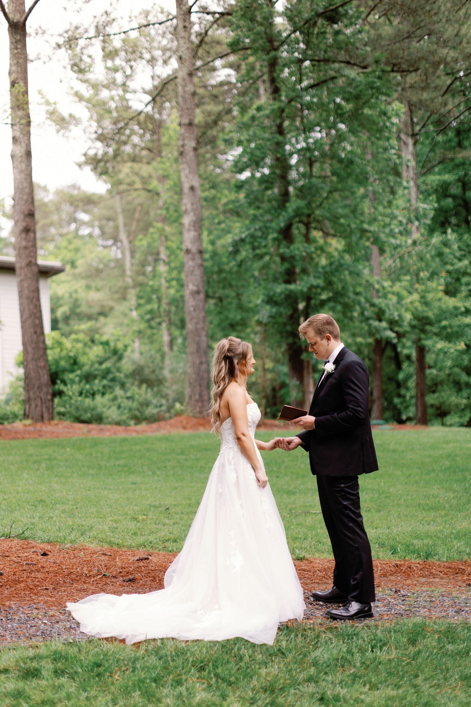 Wedding couple shares a moment on grass surrounded by tall pine trees.