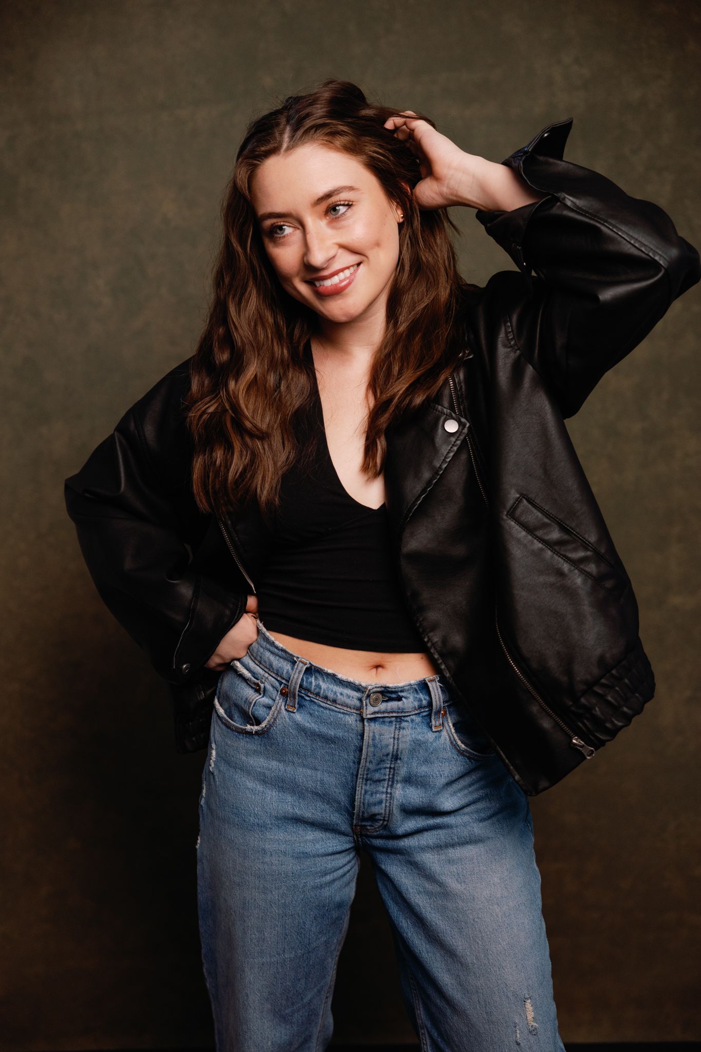 A casual portrait of someone in a black leather jacket and jeans with wavy brown hair.