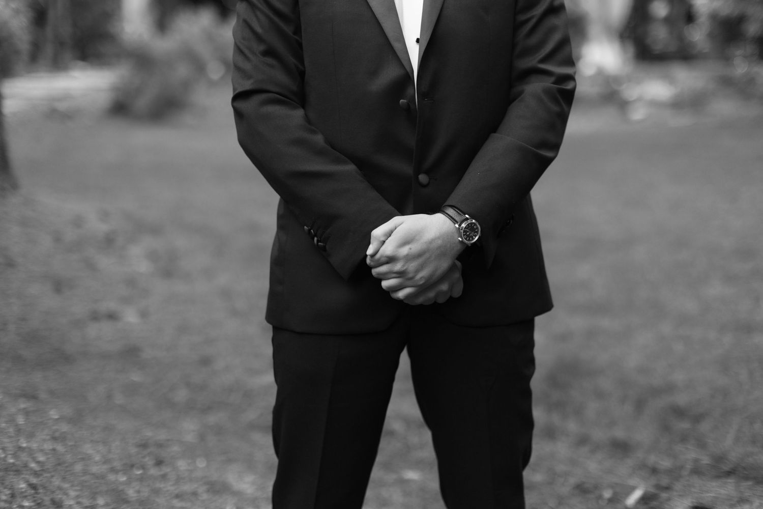 Person in formal black suit stands with clasped hands in an outdoor garden setting.