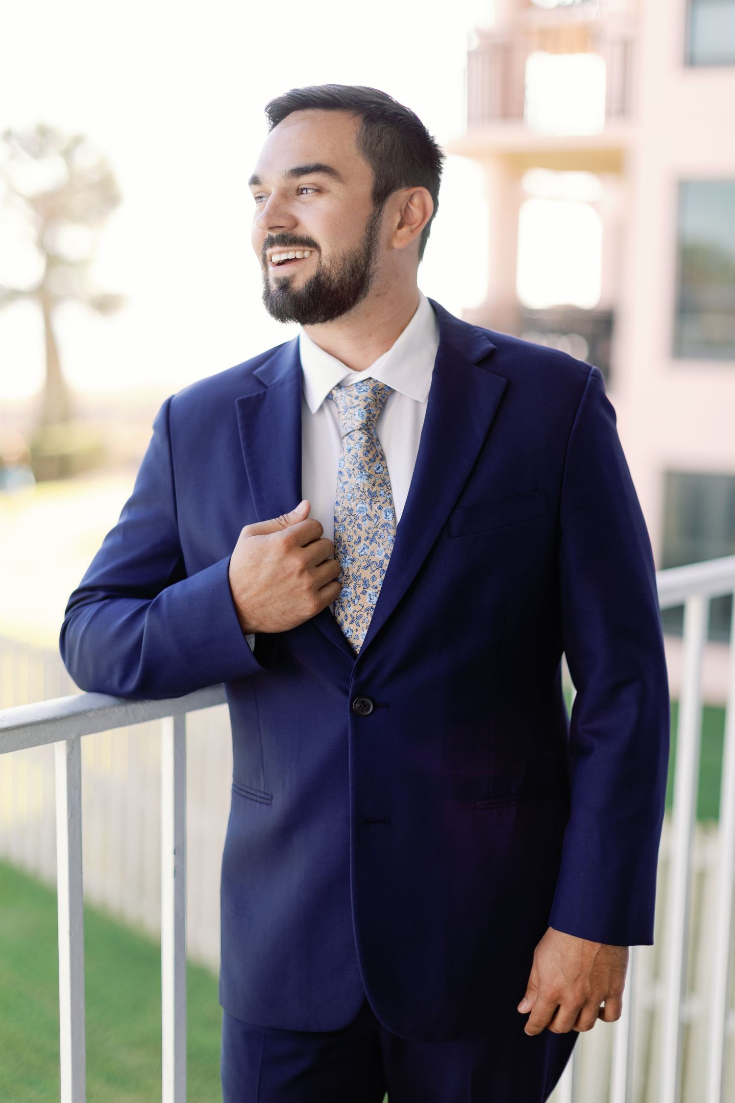A groom in a navy suit and patterned tie adjusts his collar on a balcony.