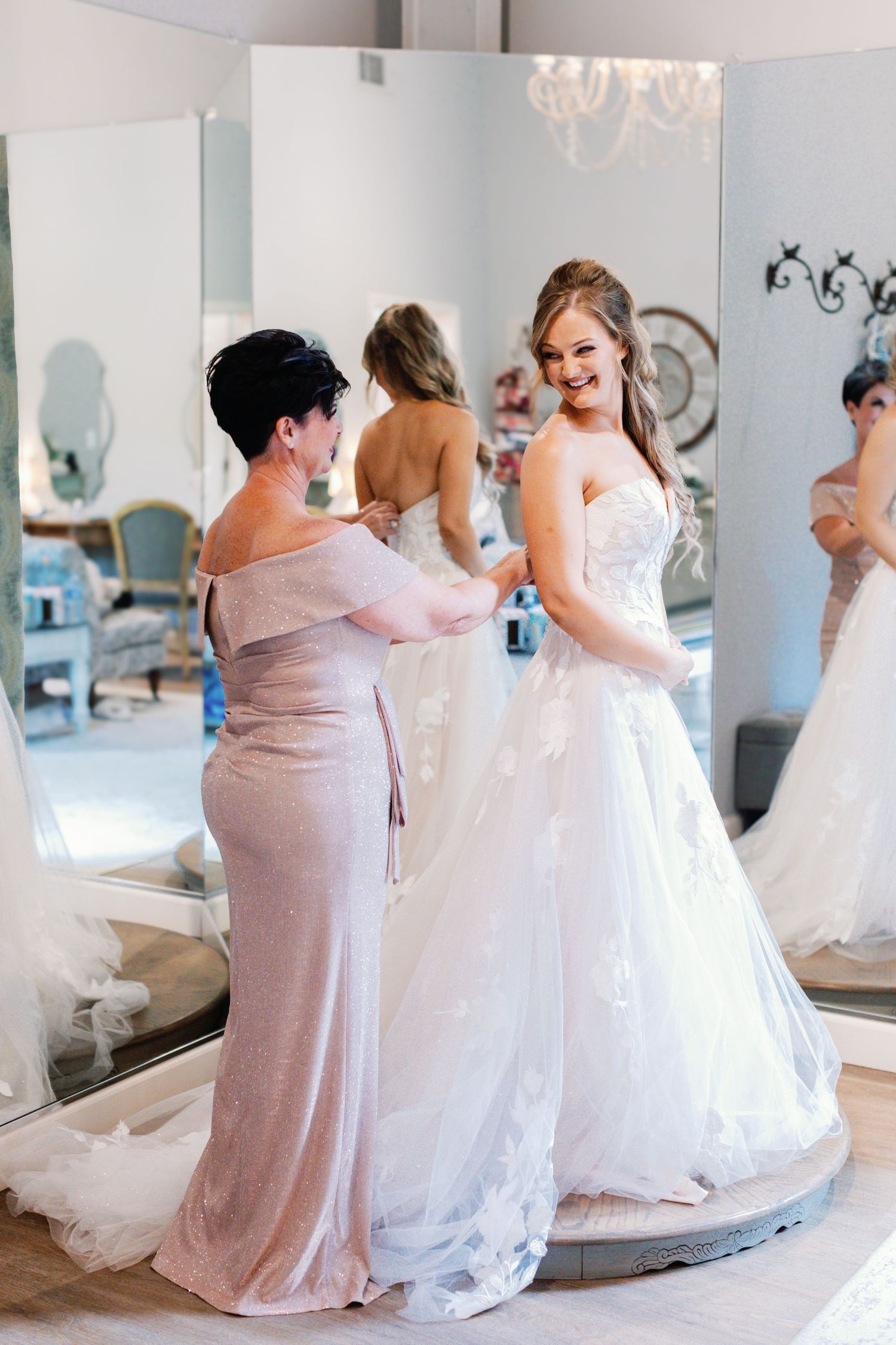 Bridesmaids in mauve dresses help adjust the bride's gown in a mirrored room.
