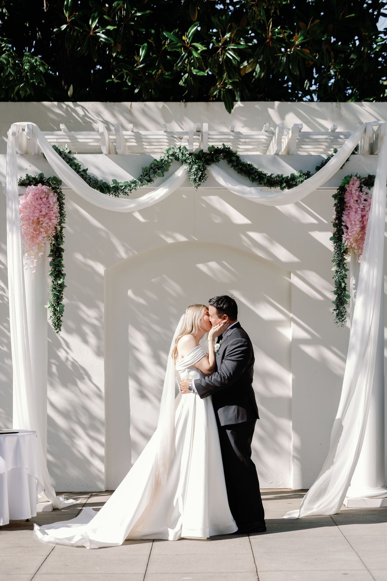 Romantic wedding couple shares tender moments against a white draped wall with greenery decorations.