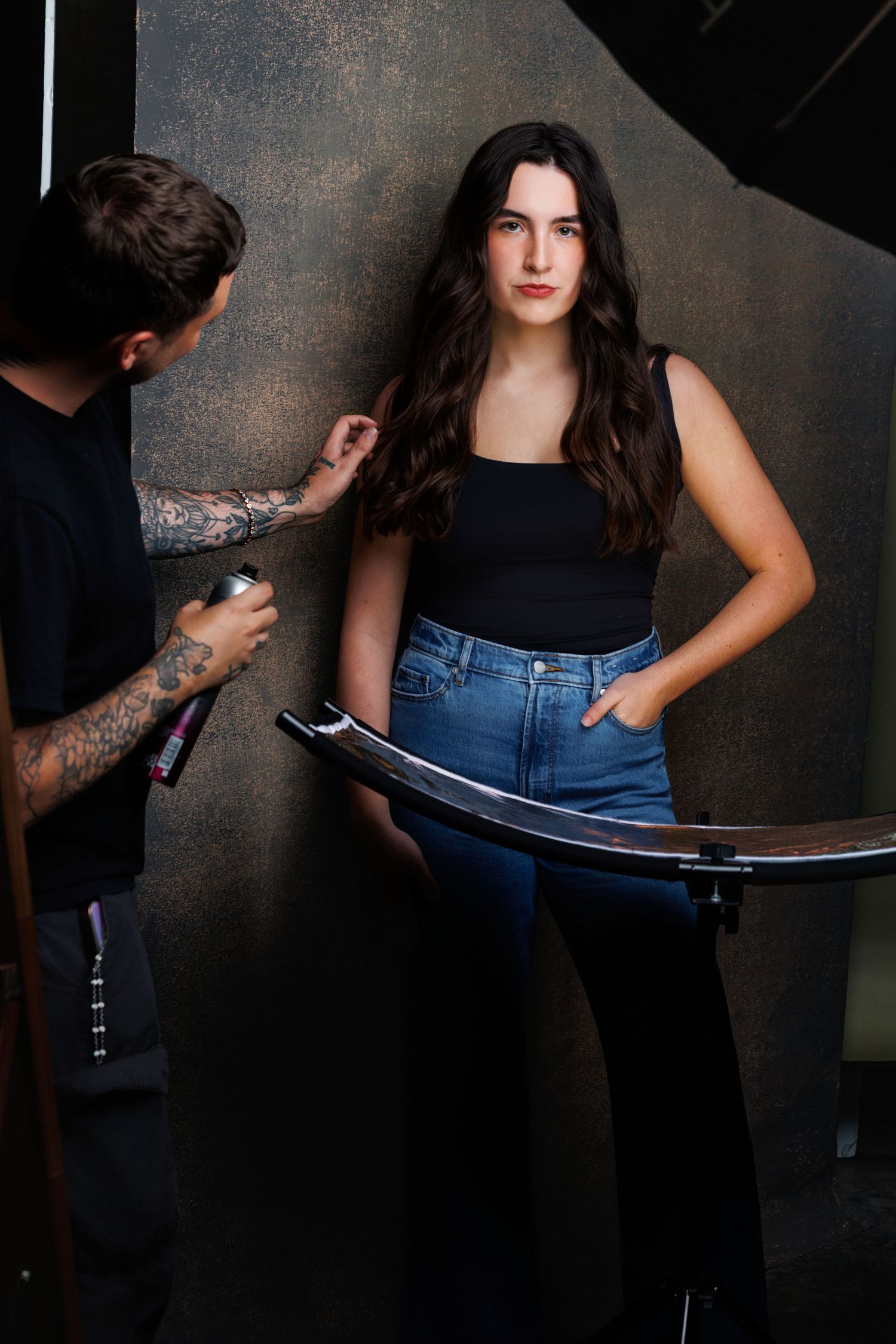 Fashion photoshoot series showing model in black tank top and jeans being photographed against dark textured backdrop.