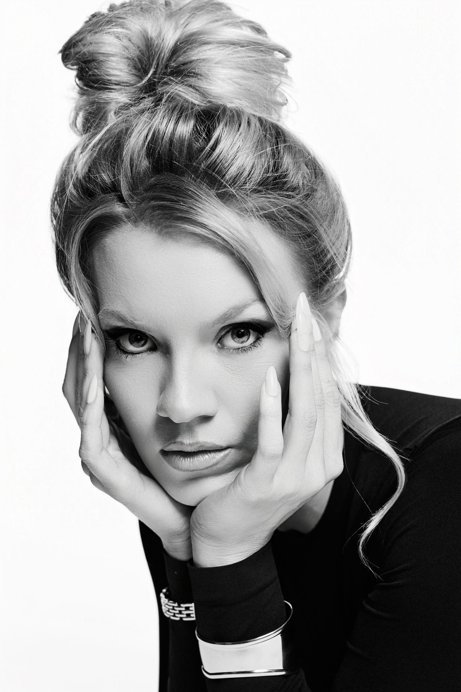 Black and white portrait of a blonde woman with an updo hairstyle, hands framing her face, wearing a dark top.