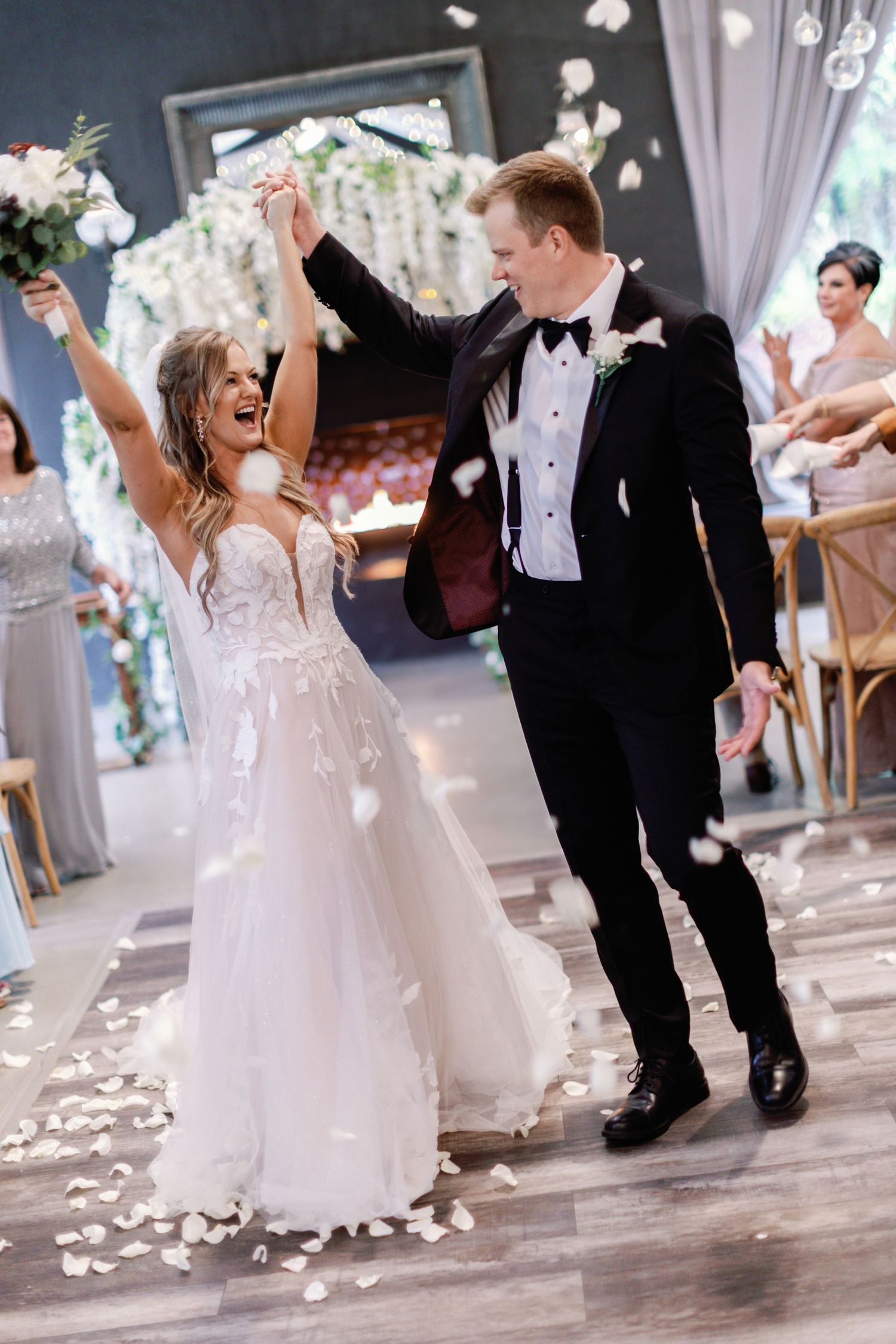 Newlyweds dance joyfully while white flower petals fall around them on their wedding day.