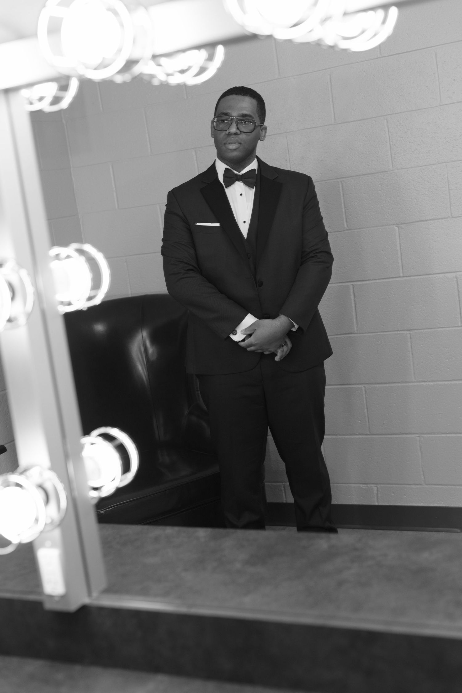Groom in classic black tuxedo poses thoughtfully against white wall with studio lighting.