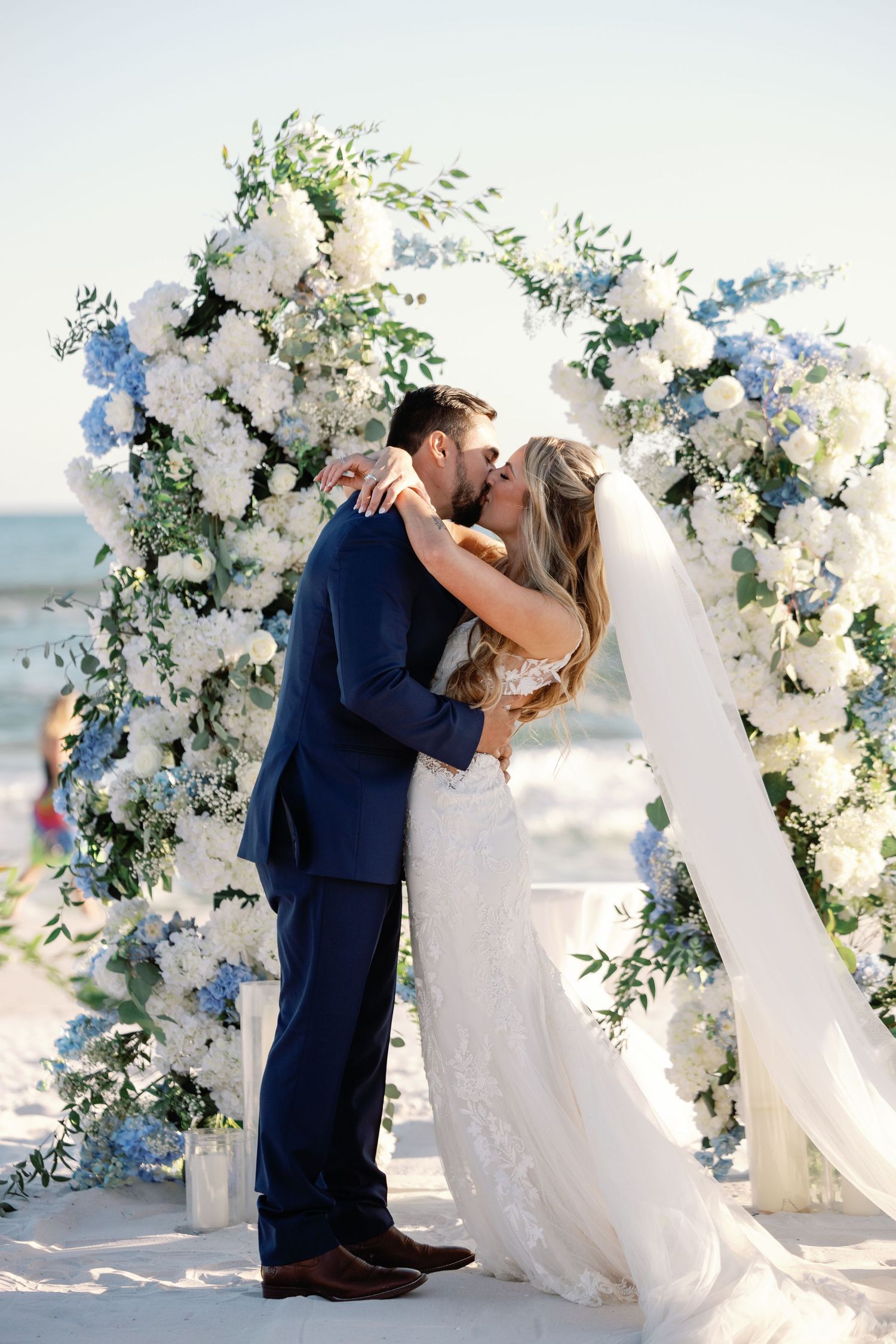 Newlyweds share their first kiss under a floral arch on a sunny beach.
