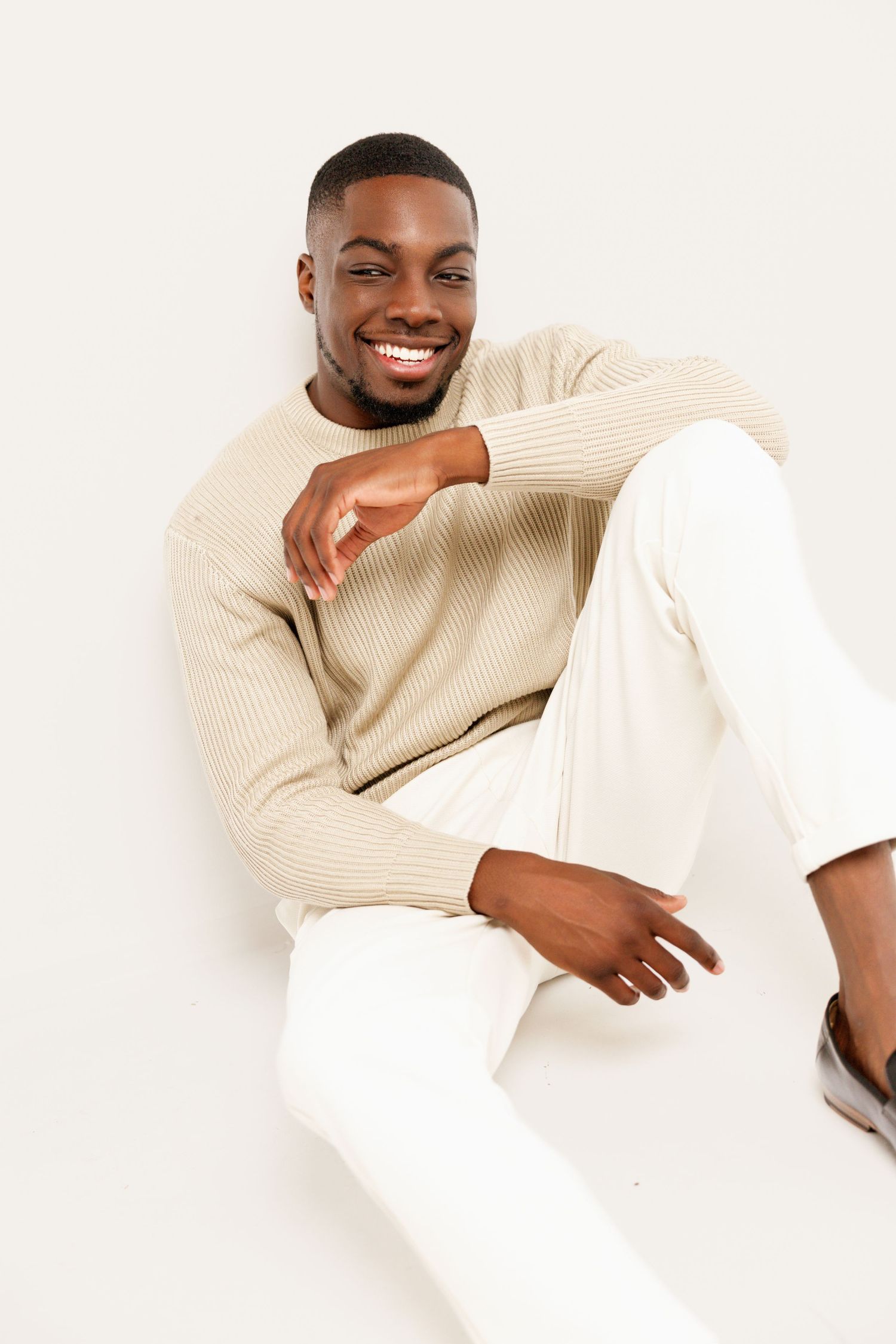 A relaxed portrait of someone in cream knitwear and white pants sitting casually.