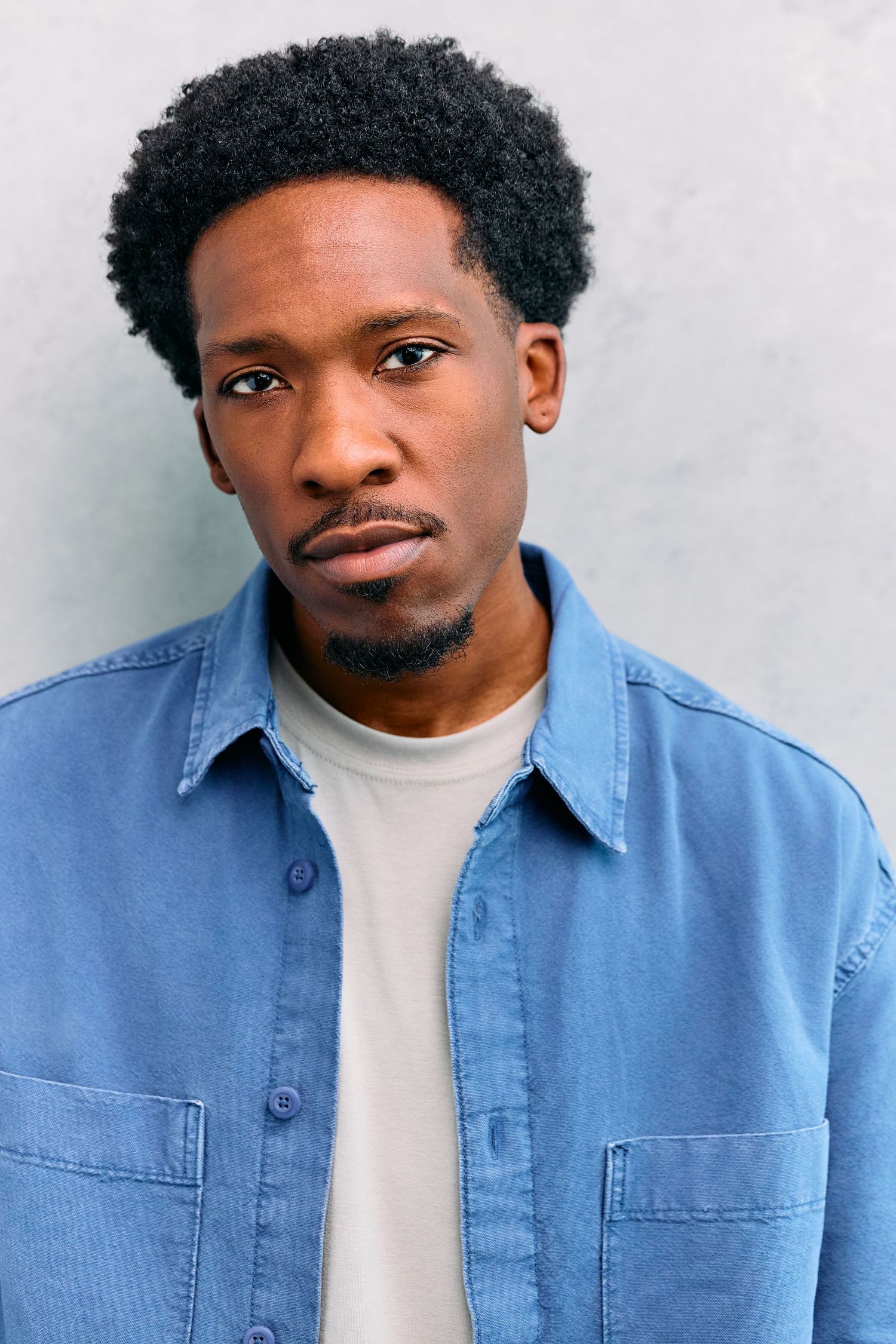Young Black man with afro hair wearing a blue denim shirt over white tee, posing against a gray wall.