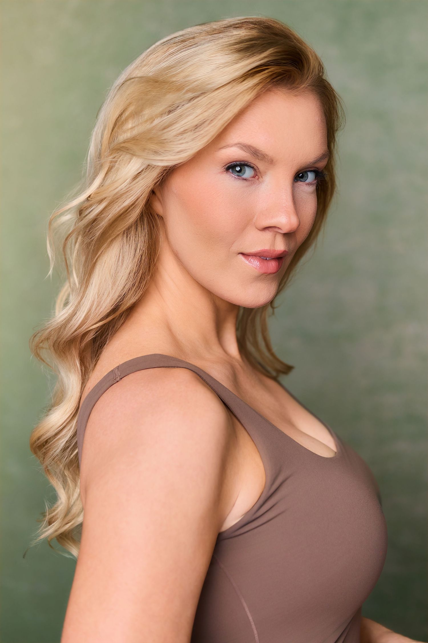 Blonde woman with blue eyes wearing a brown tank top poses against a green background.