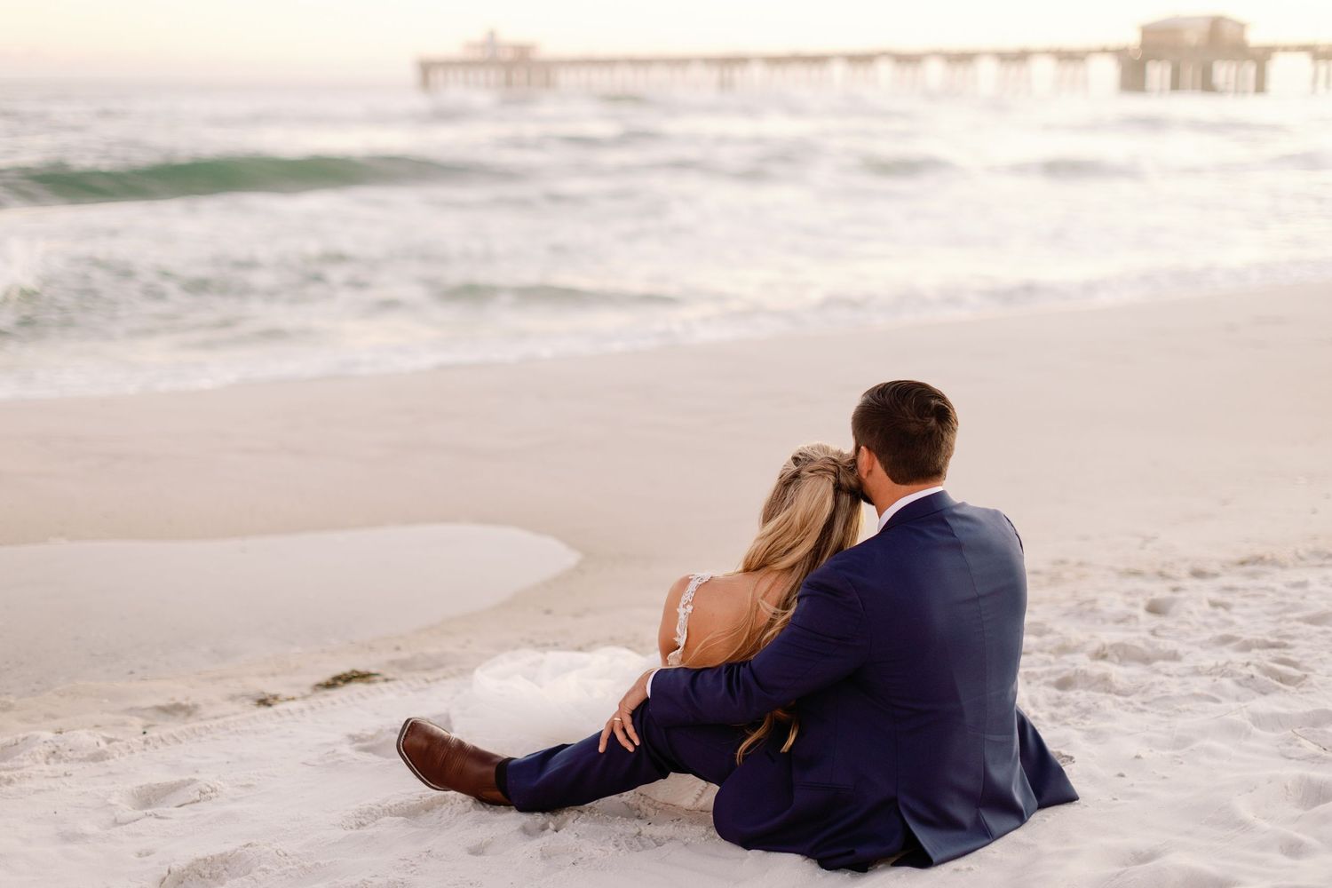 Couple sits together on beach at sunset with waves and buildings in background.