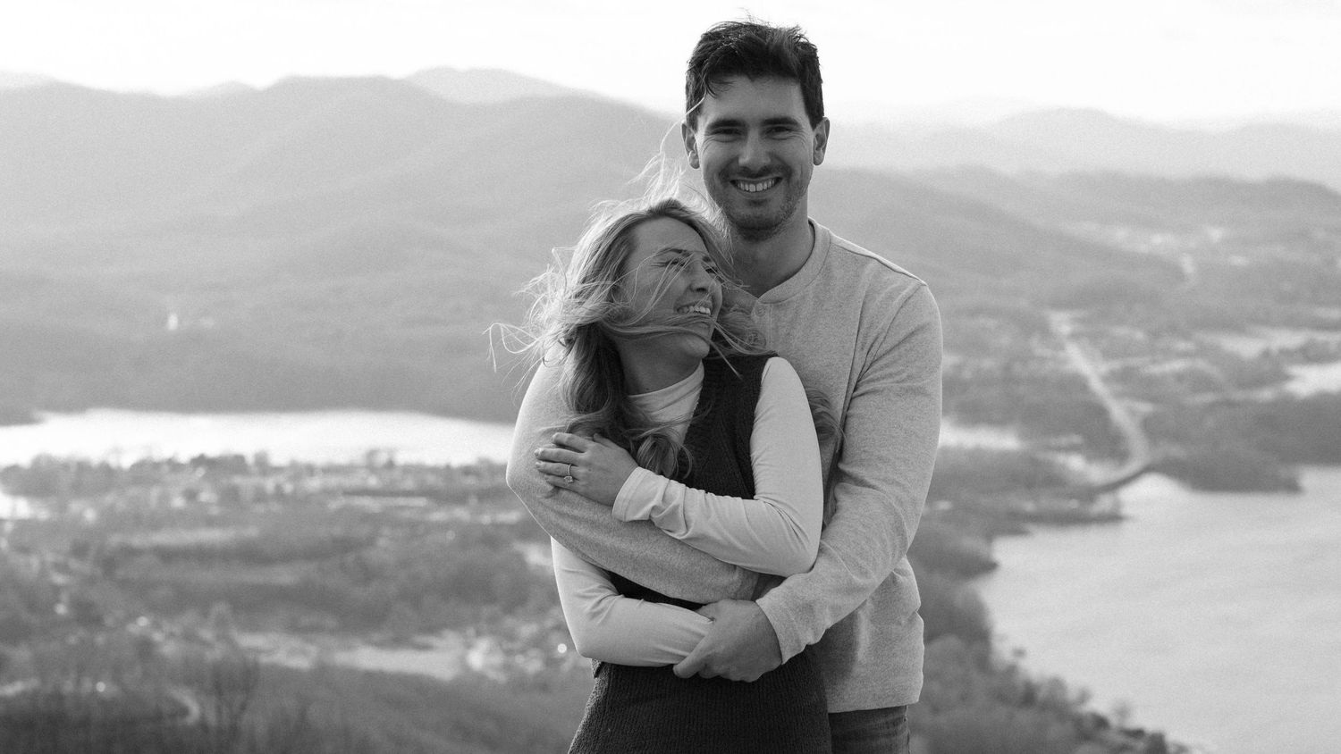 Panoramic black and white shot of a couple embracing on a mountain vista overlooking water.