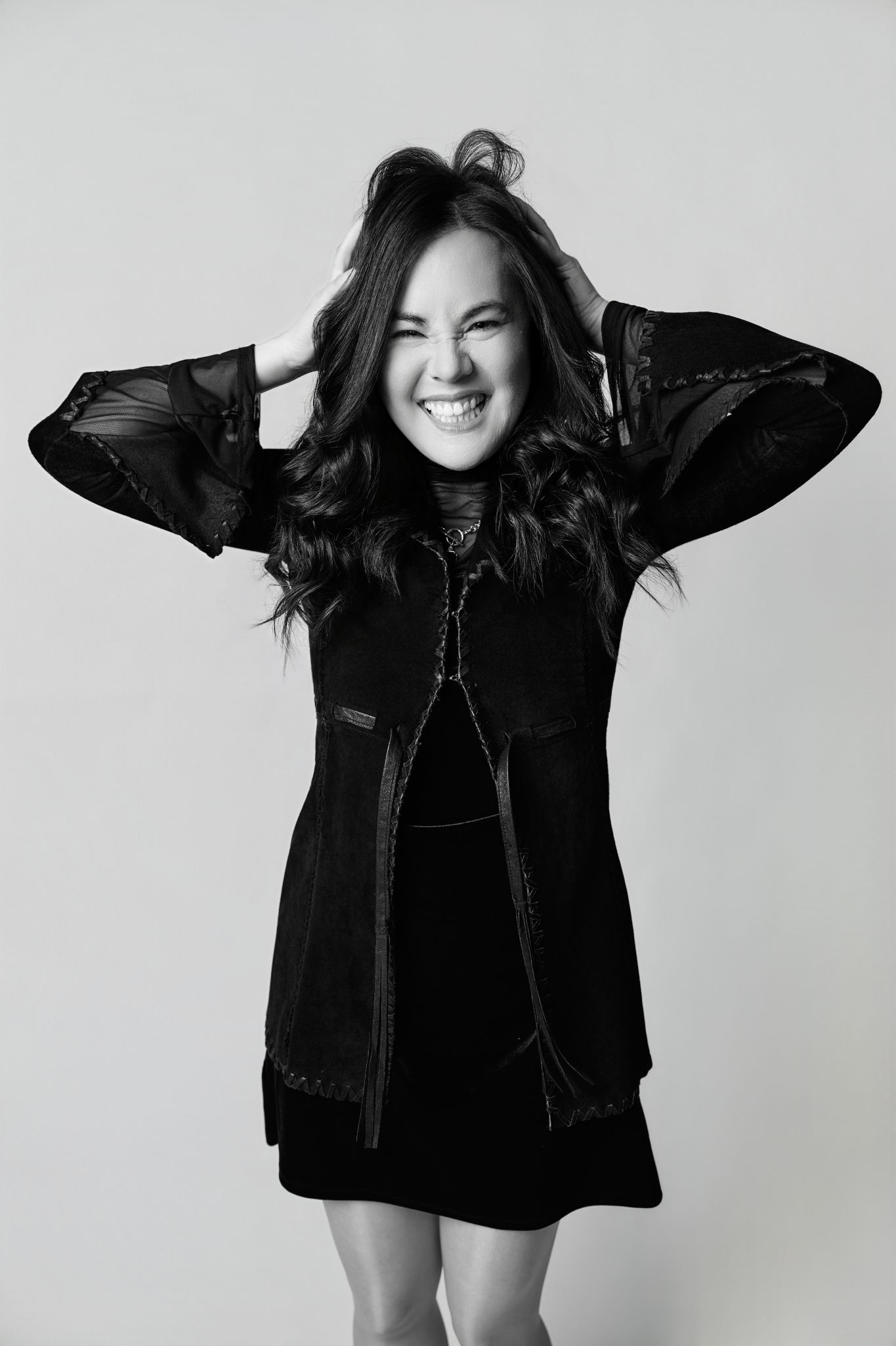 Black and white photo of a smiling young woman in a black dress, hands in hair, posing against a light background.
