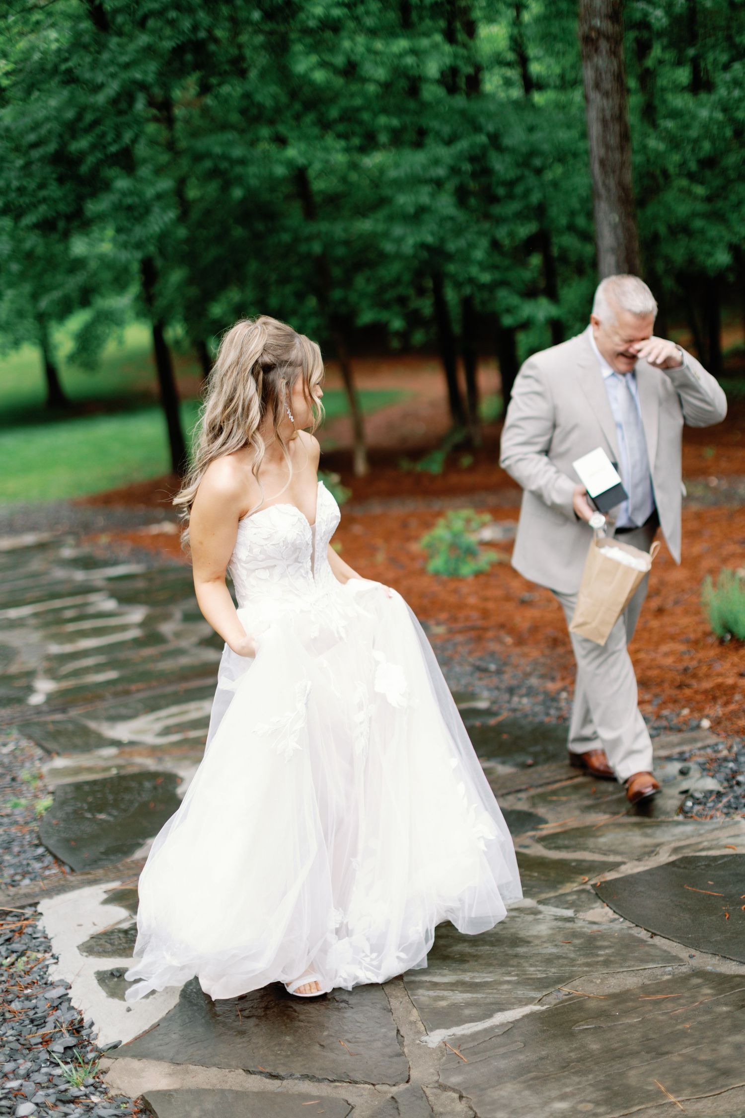 A bride in a white gown walks along a stone path while someone in gray follows behind.