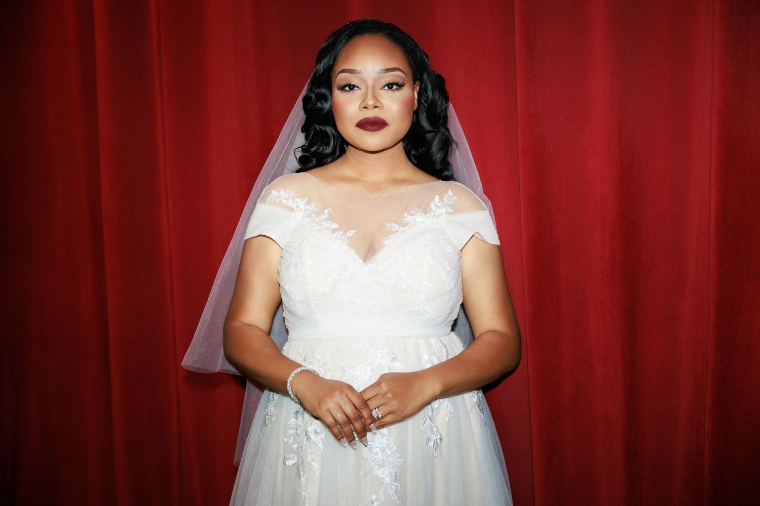 Bride in elegant lace wedding dress poses against red velvet curtain backdrop.