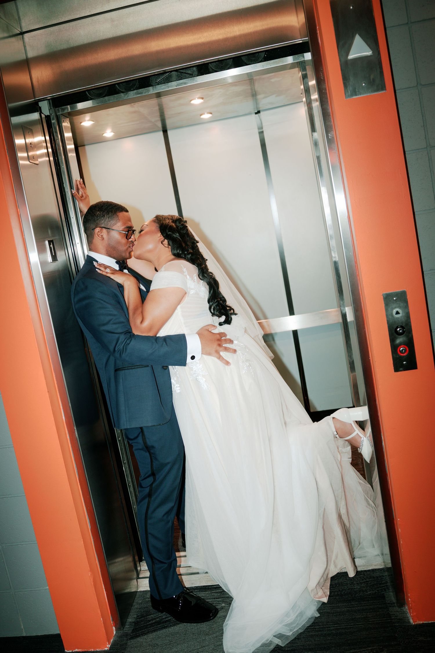A romantic moment between newlyweds sharing a kiss in an elevator with orange walls.