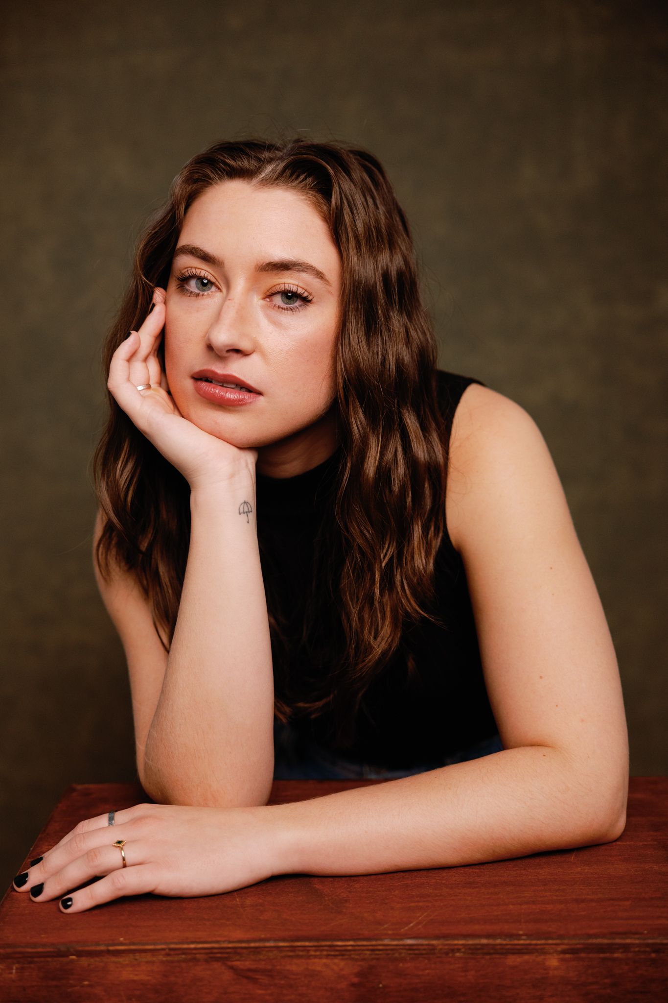 A studio portrait of someone in black top resting their head on their hand at a wooden table.