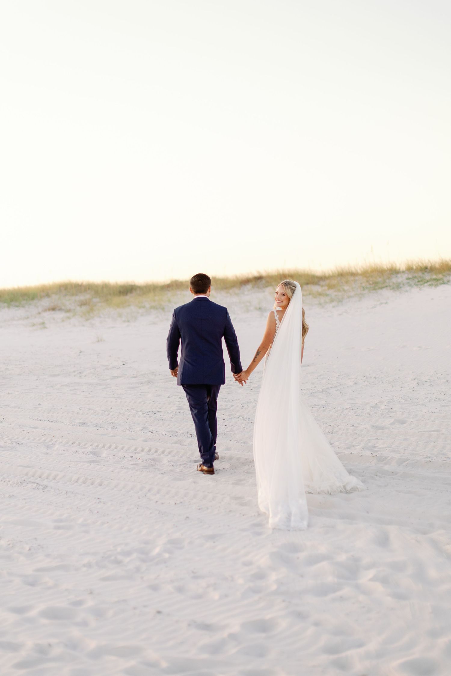Newlyweds walk hand in hand across white sand dunes at beach wedding.