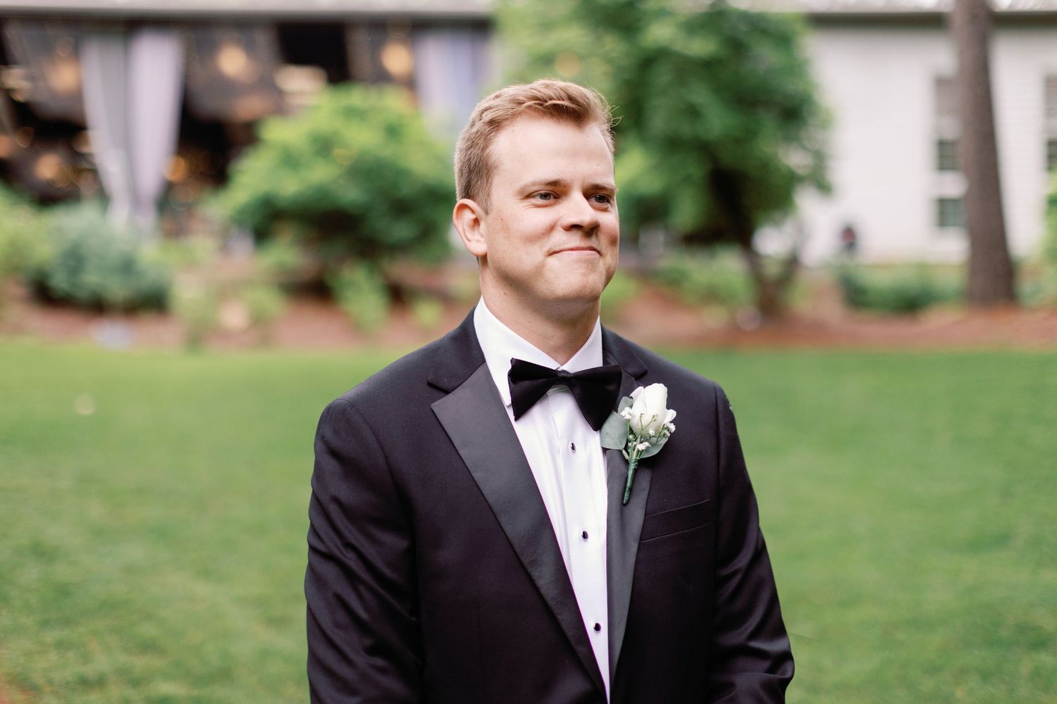 A man in formal attire stands outdoors in a garden setting wearing a black tuxedo and boutonniere.
