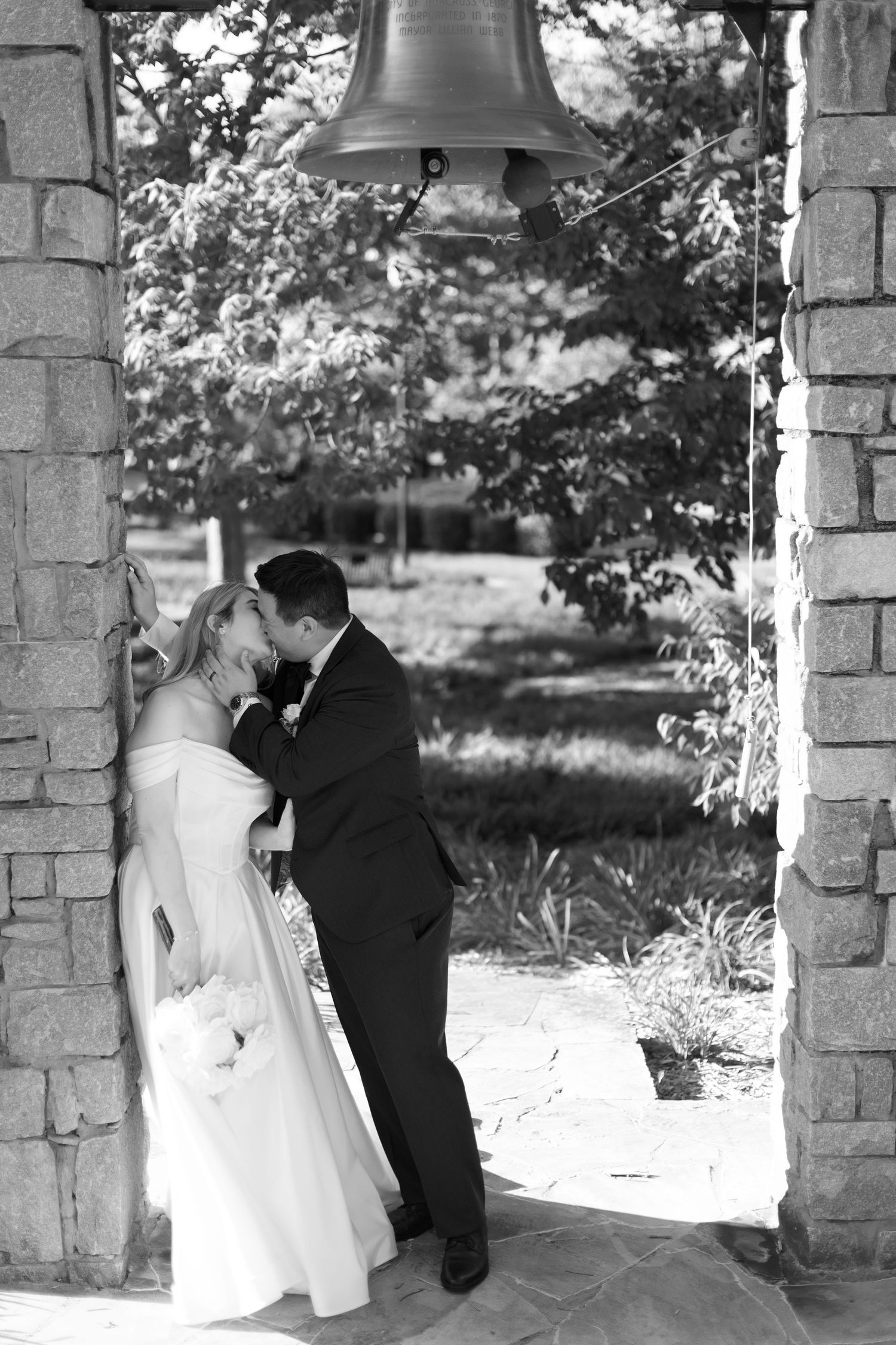 Newlyweds share an intimate moment under a stone bell arch in black and white photography.