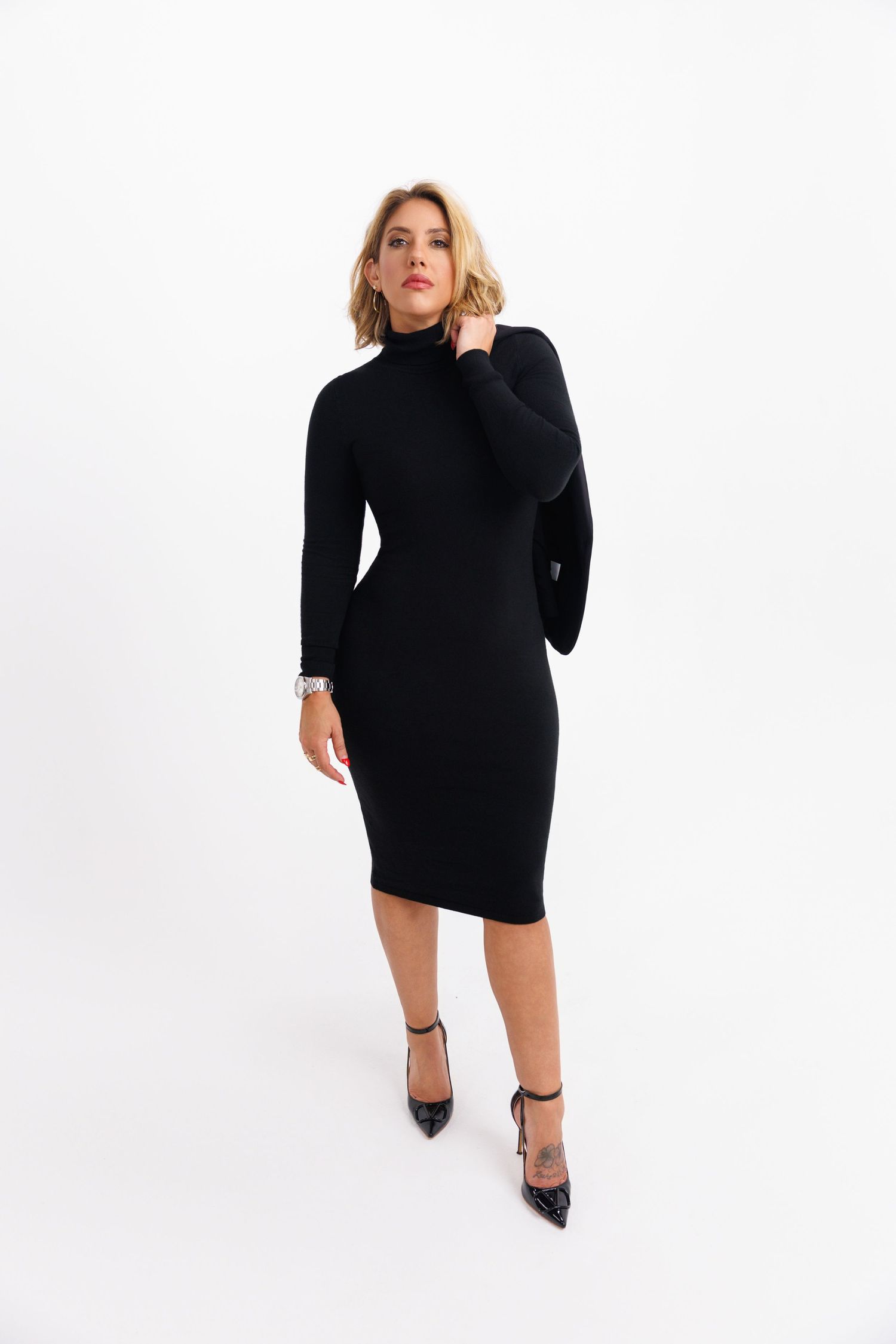Woman in a short black turtleneck dress and stiletto heels poses confidently against a white backdrop.