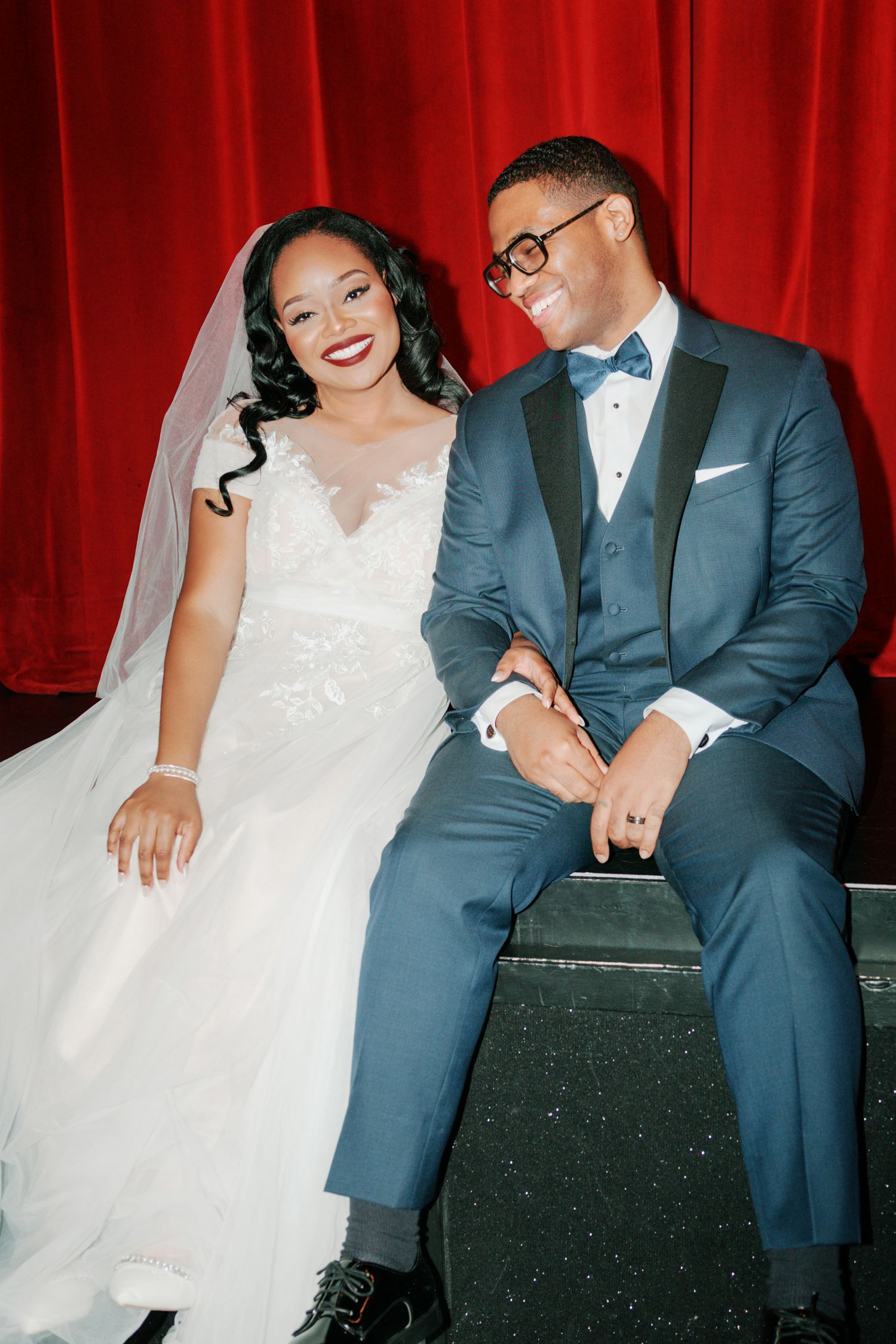 Wedding couple sitting together on stage against vibrant red theater curtain.