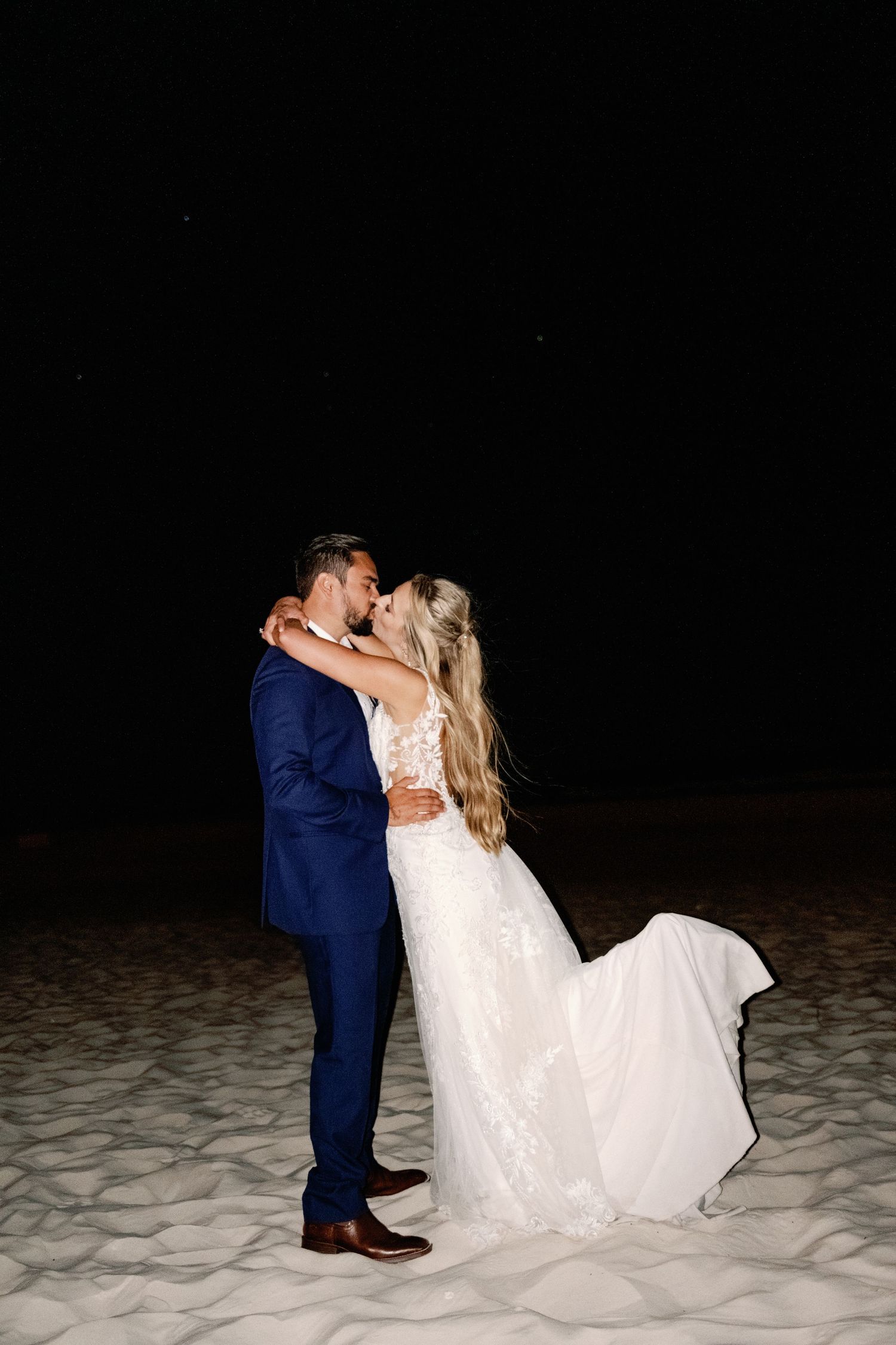 Romantic beach photoshoot of couple in wedding attire against dark background.