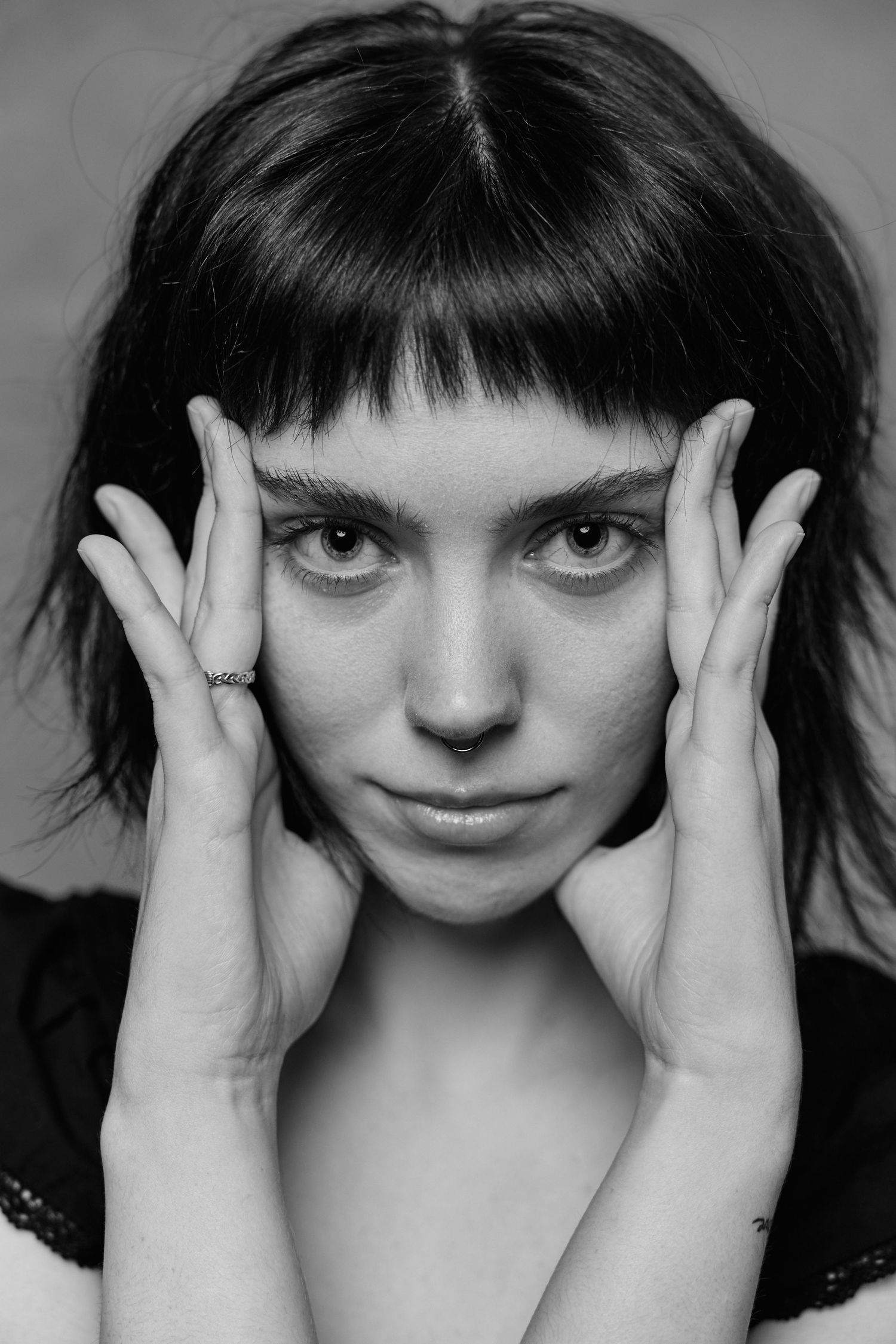 Black and white portrait of a young woman with bangs holding her face in her hands, wearing a ring.
