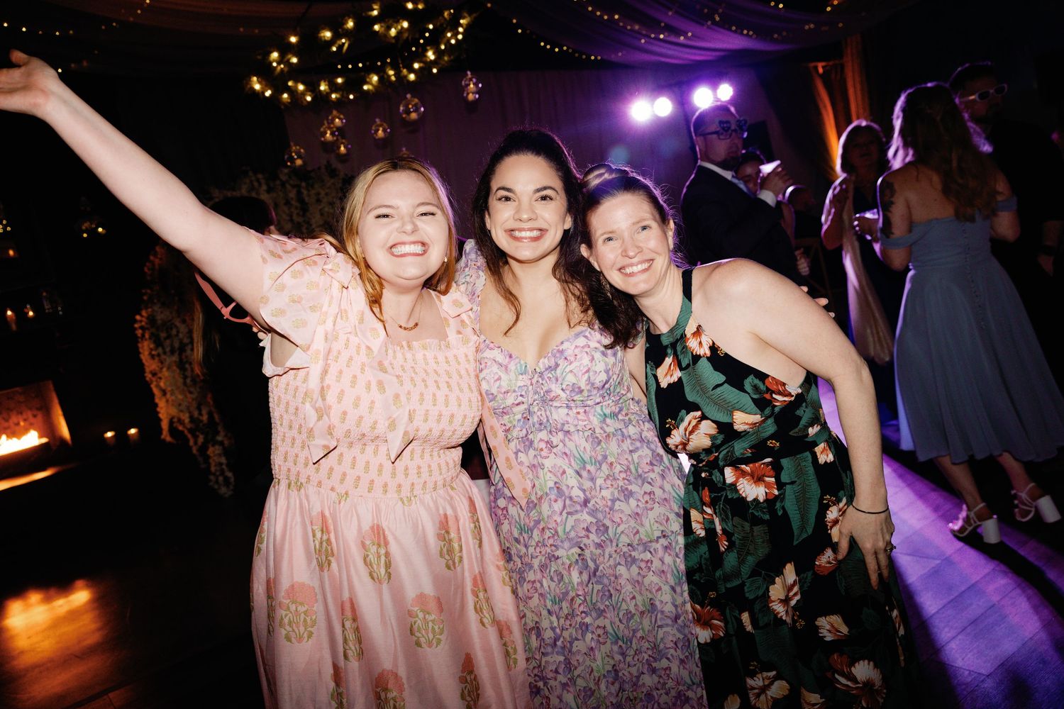 Three friends celebrate joyfully on a dance floor decorated with string lights.