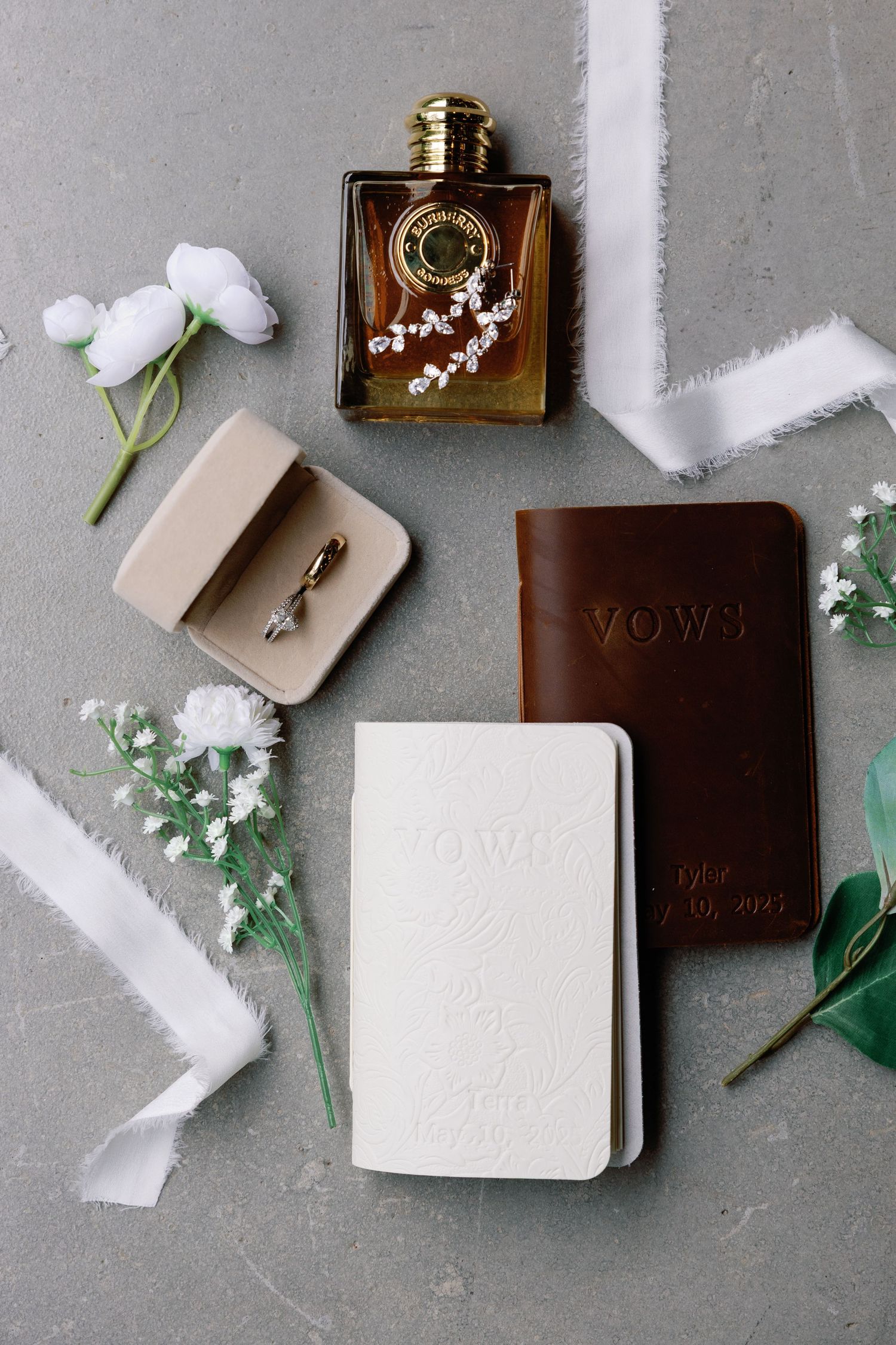 Wedding details flatlay with vow books, perfume, rings, and white flowers on grey background.