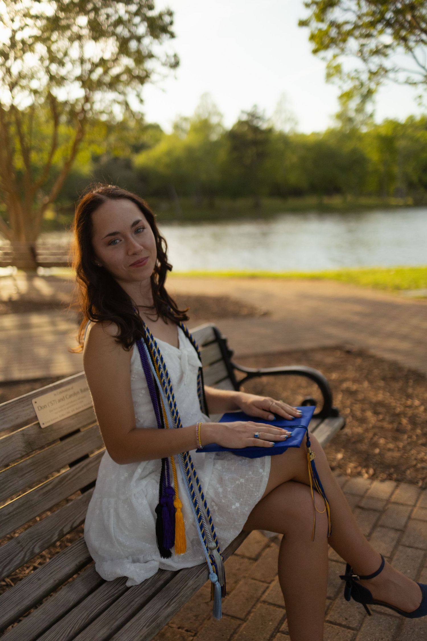 Graduate sitting on wooden bench near waterfront at sunset.