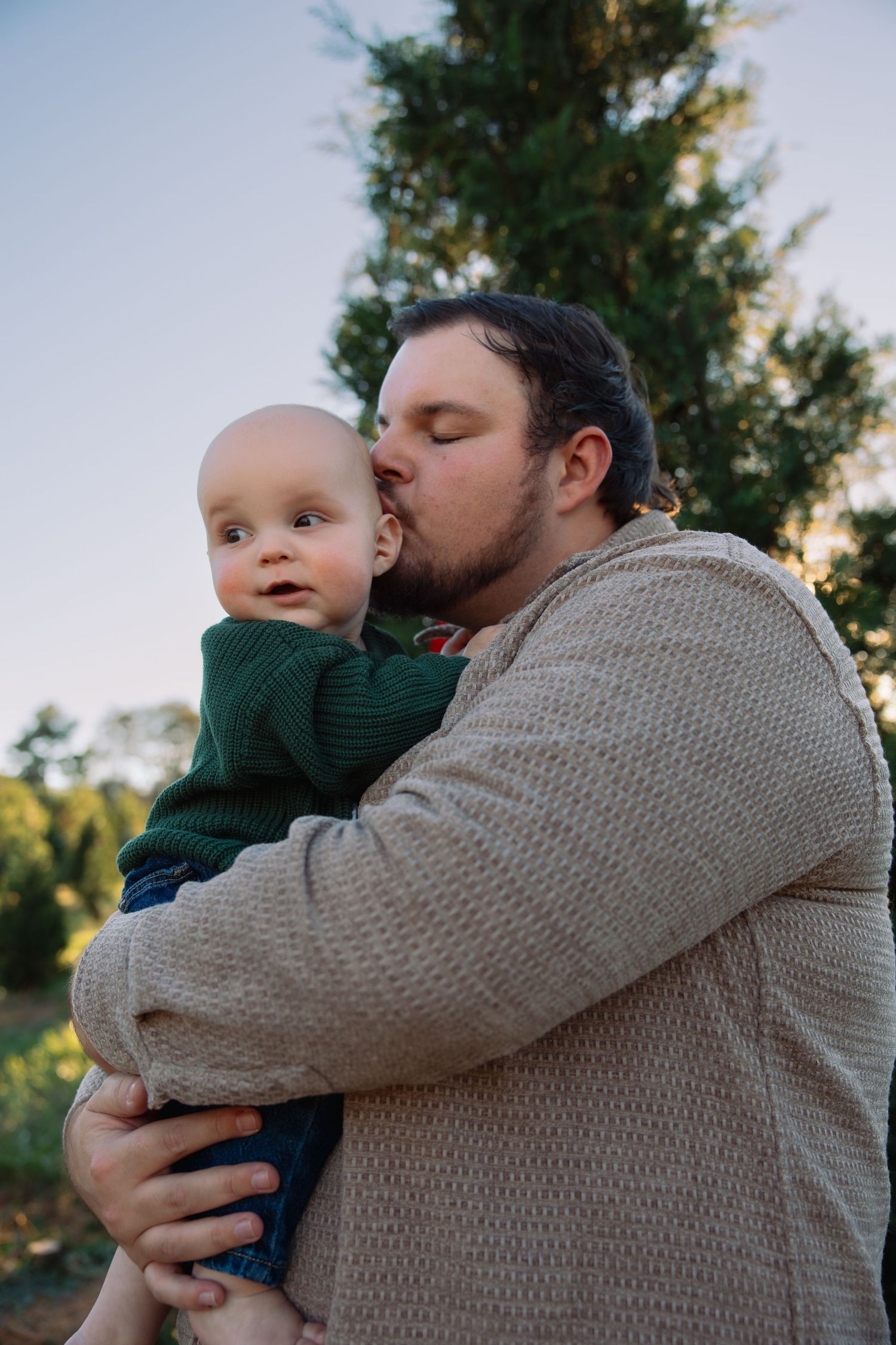 Tender moment shared between parent and child against evergreen tree at sunset.