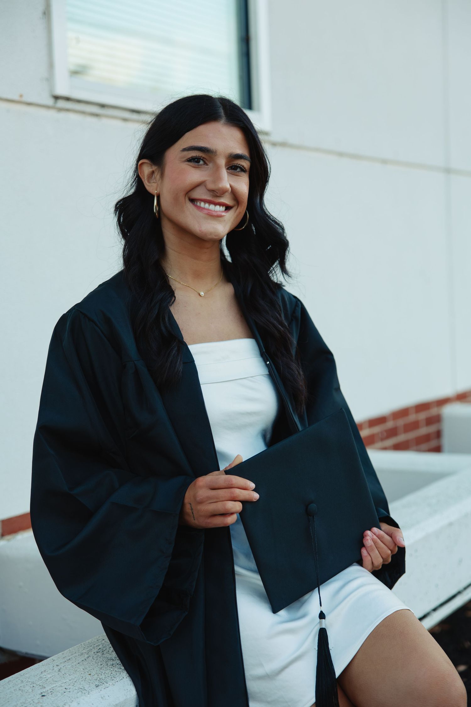 Graduate in black cap and gown poses against white wall during graduation photoshoot.