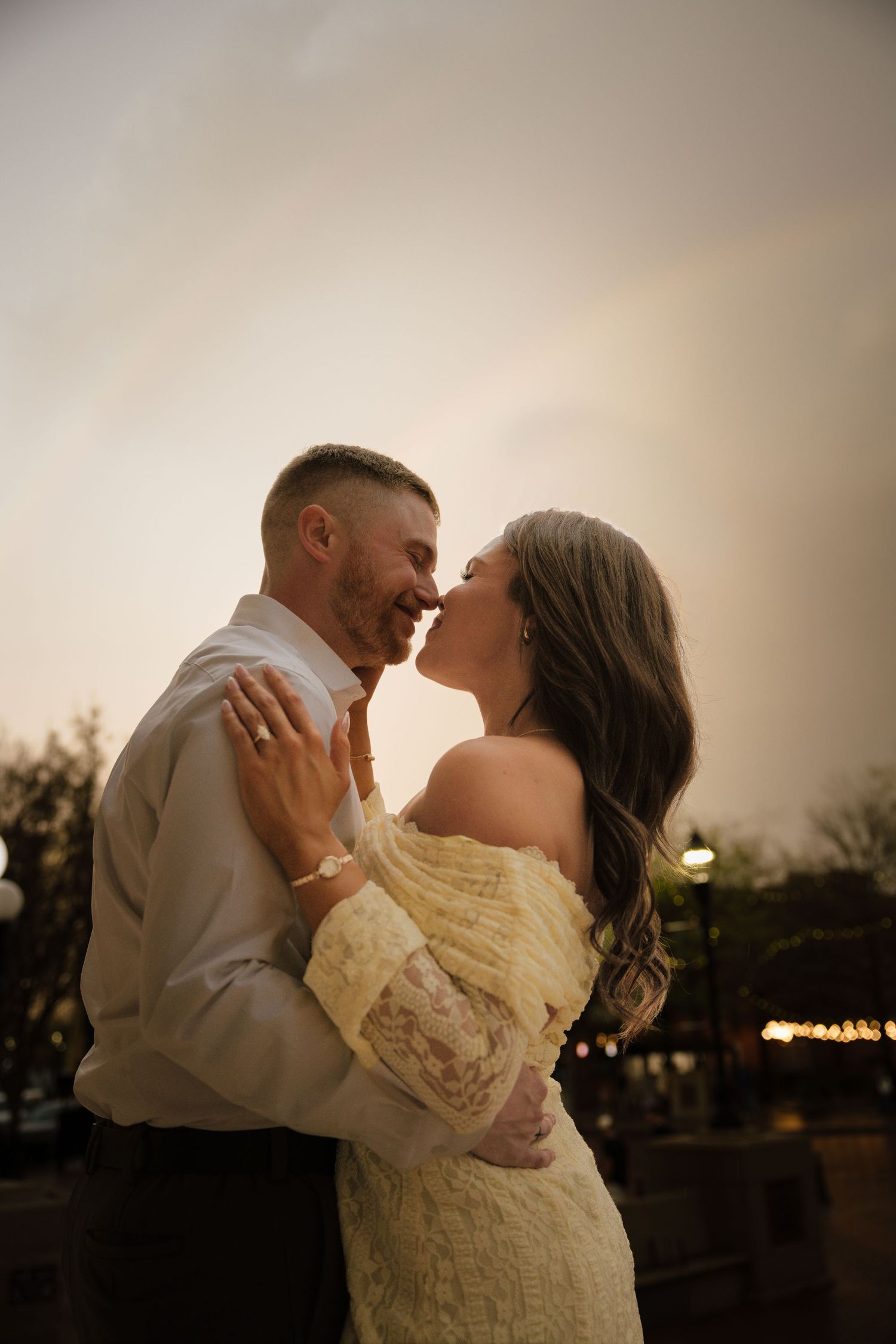 Romantic couple sharing an intimate moment at dusk, woman in yellow lace off-shoulder dress, man in beige shirt, smiling.
