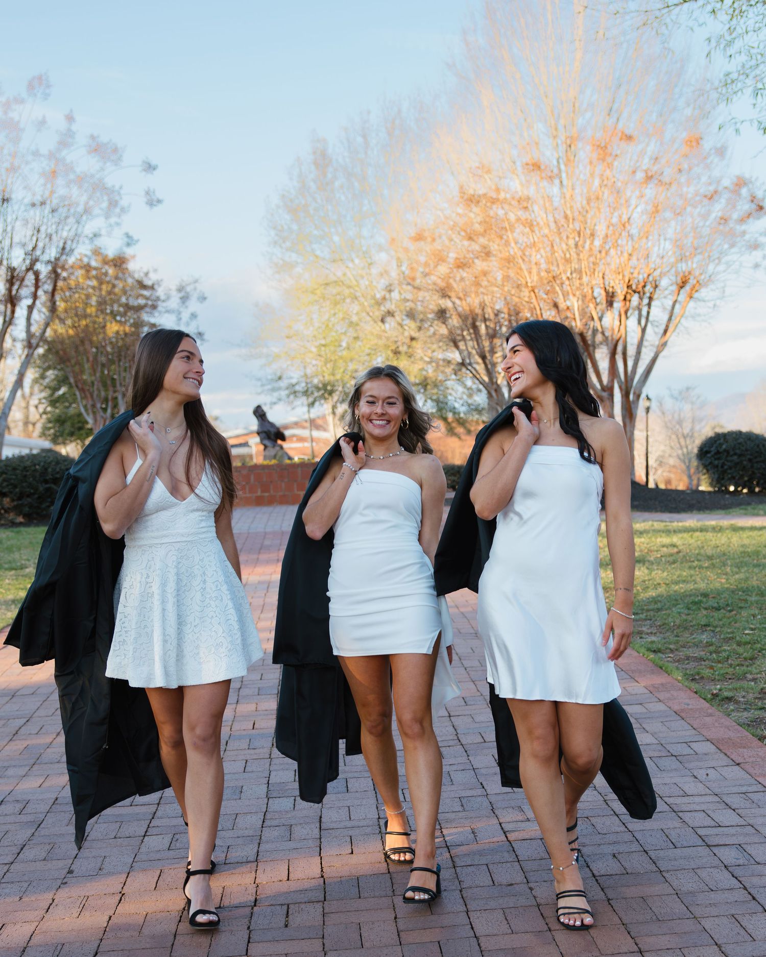 Four people in white dresses and black jackets laugh and pose together on a brick pathway at sunset.