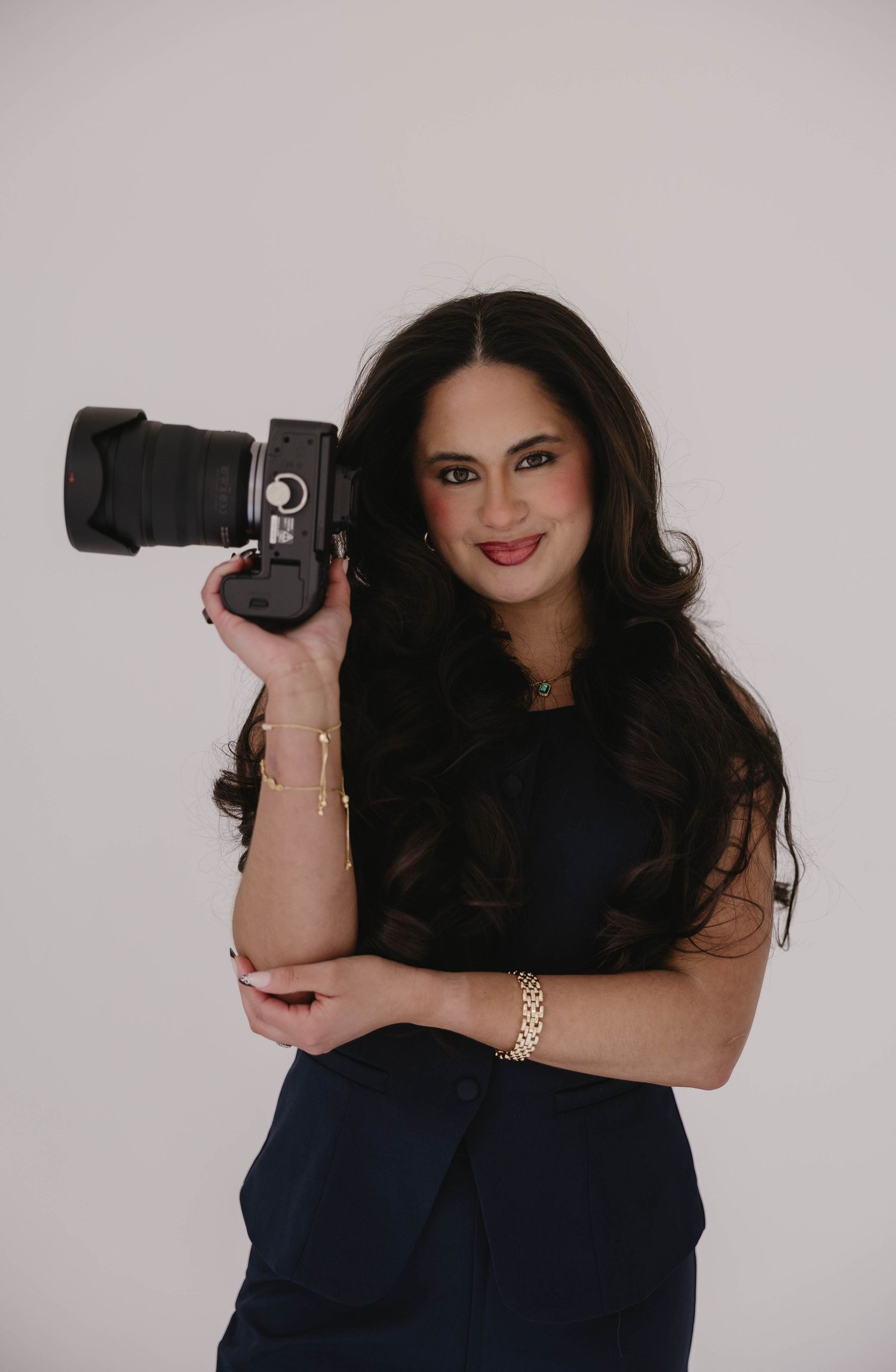 A smiling woman with long dark hair holds a DSLR camera, wearing a navy dress and gold jewelry against a white background.