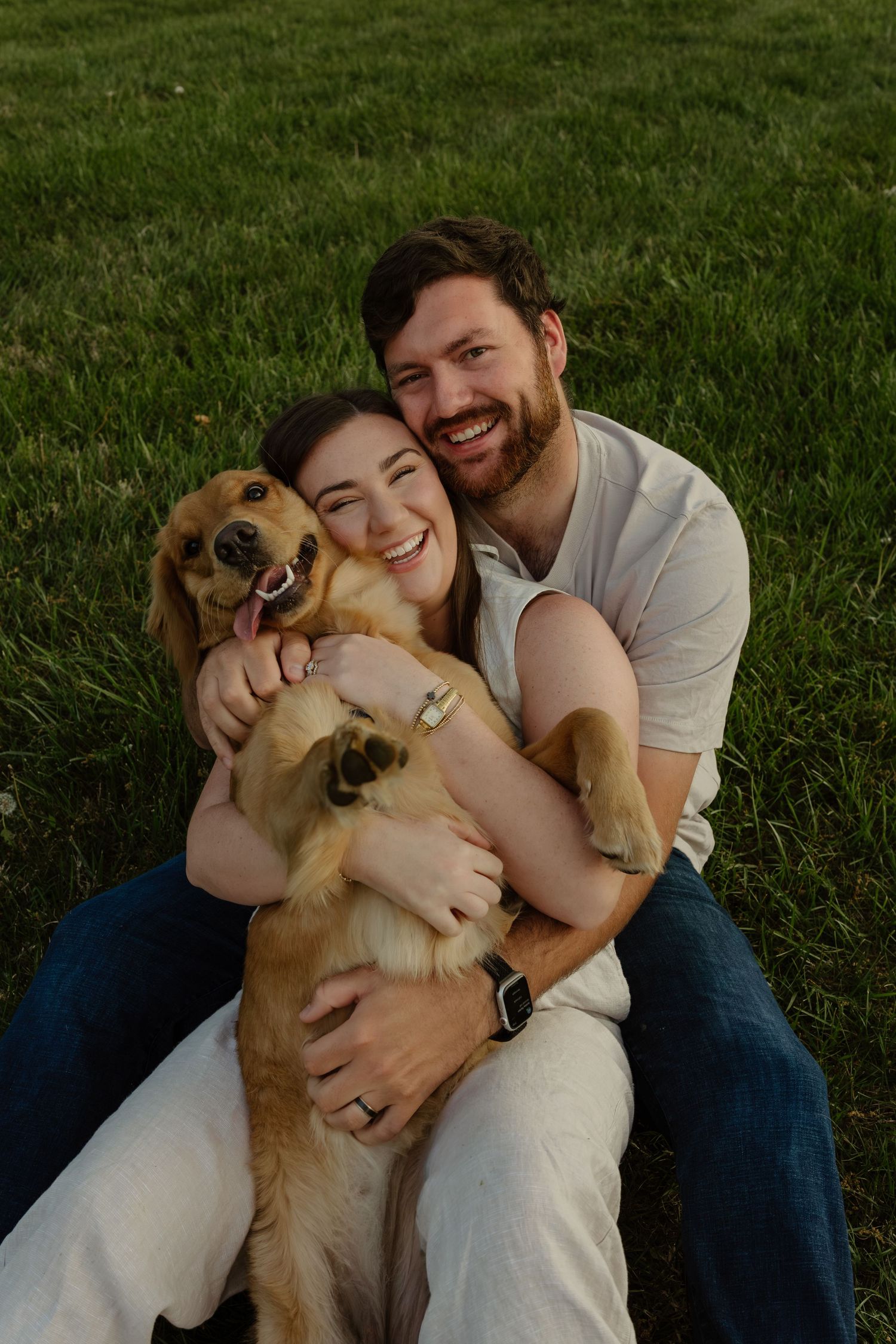 Happy couple sitting on green grass, smiling and hugging their golden retriever dog outdoors.