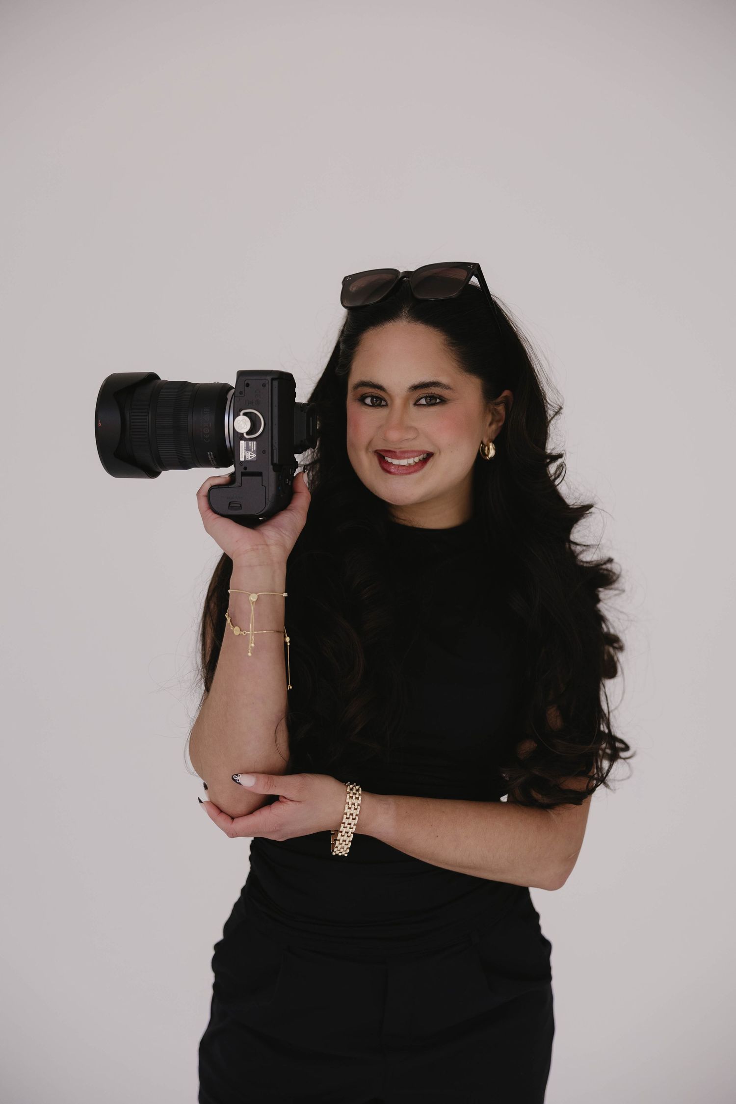 Smiling woman in black dress holding a DSLR camera with sunglasses on her head against a white background.